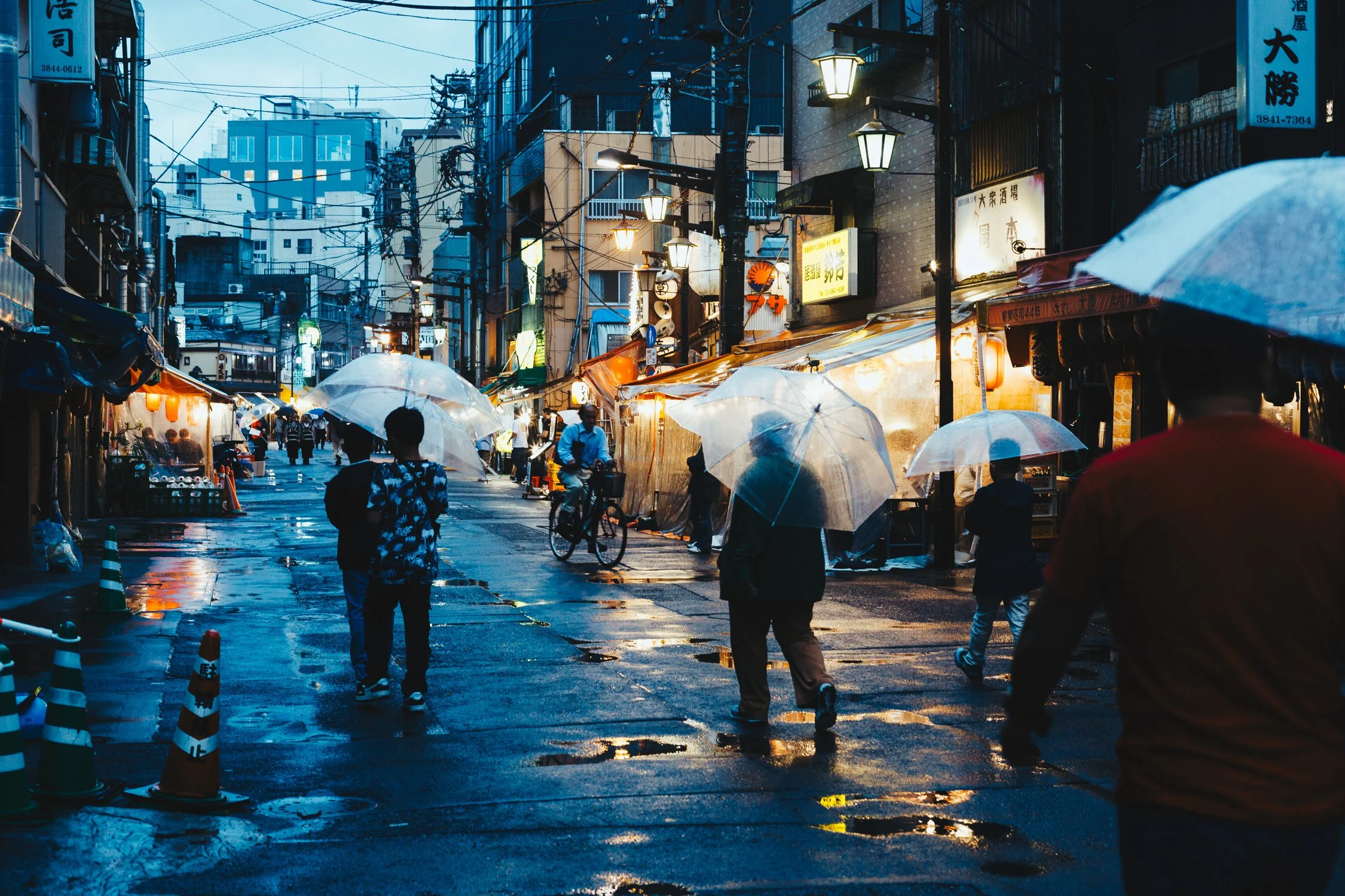 People walking and cycling on a wet street in Japan during evening rain, with umbrellas, illuminated shop signs, and traditional street lamps.