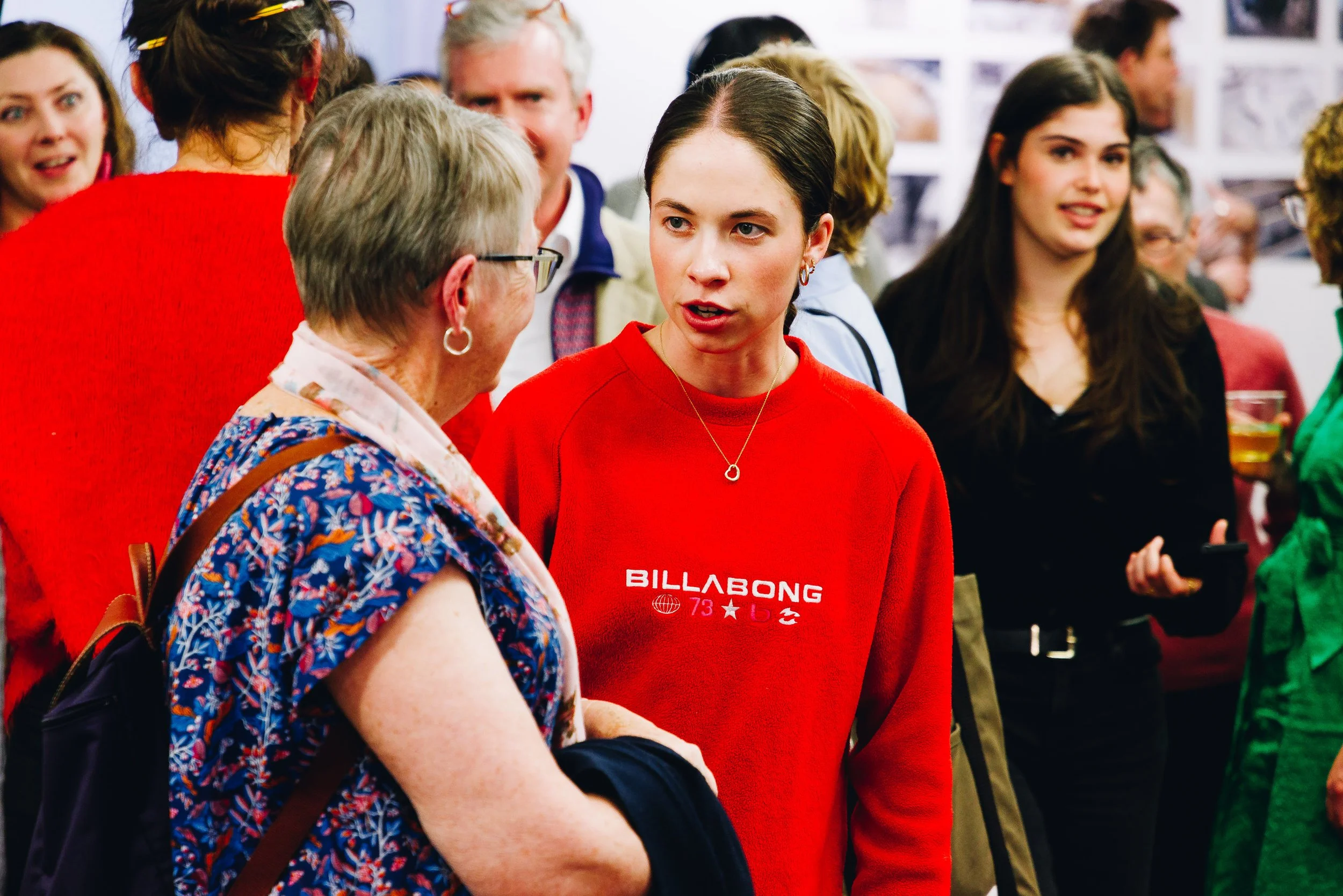 A young woman in a red sweatshirt is talking to an elderly woman with short gray hair and glasses at a crowded indoor event.