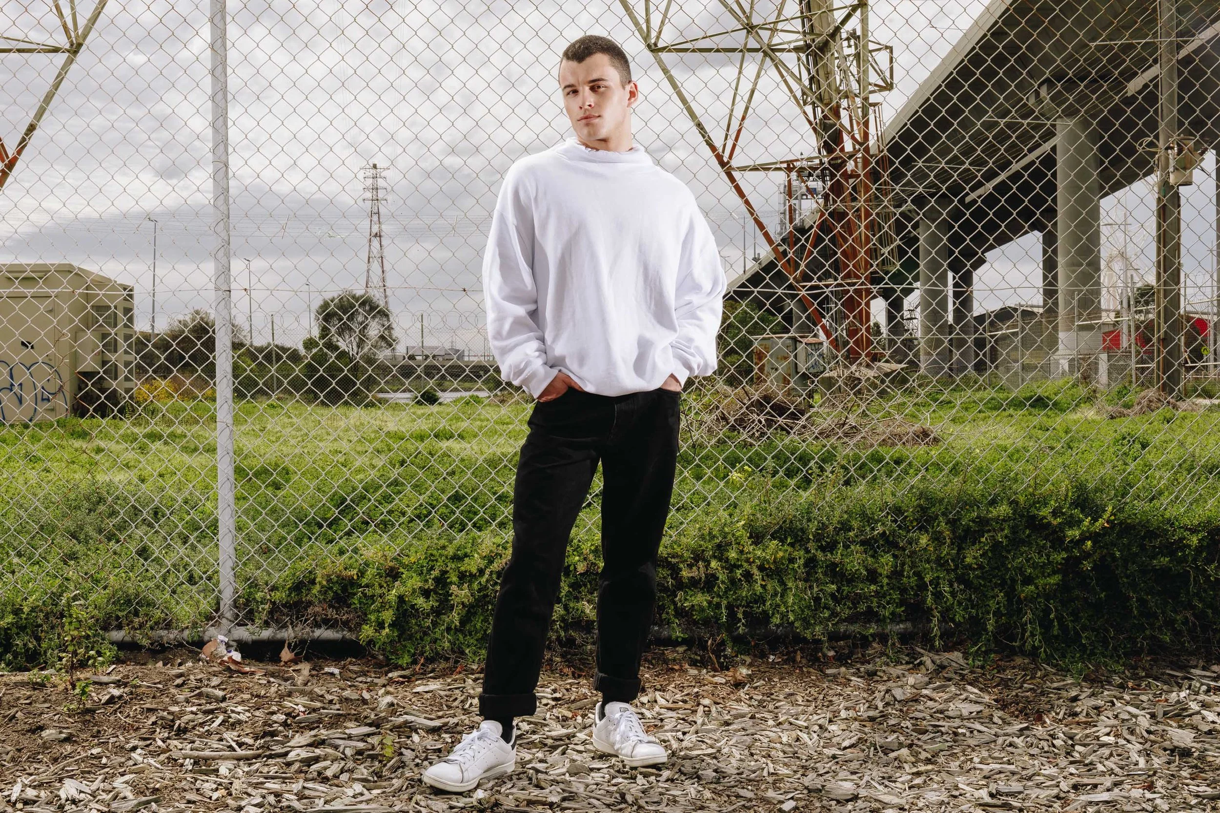A young man with short hair wearing a white oversized sweatshirt, black pants, and white sneakers stands in front of a chain-link fence on a cloudy day. Behind him, there's grass, a large metal structure, and a bridge.