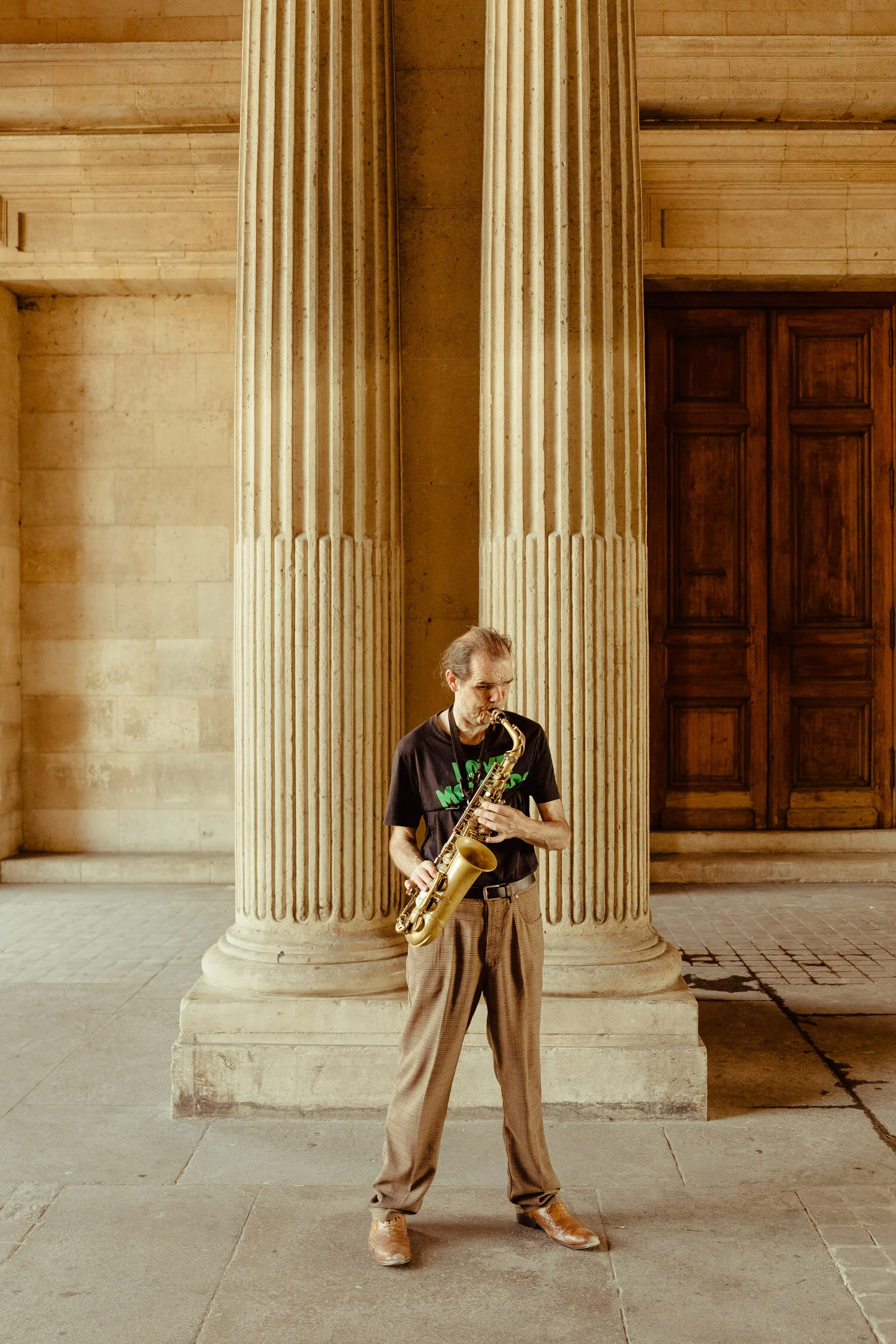 A middle-aged man playing a saxophone in front of a large classical architectural column and wooden door.