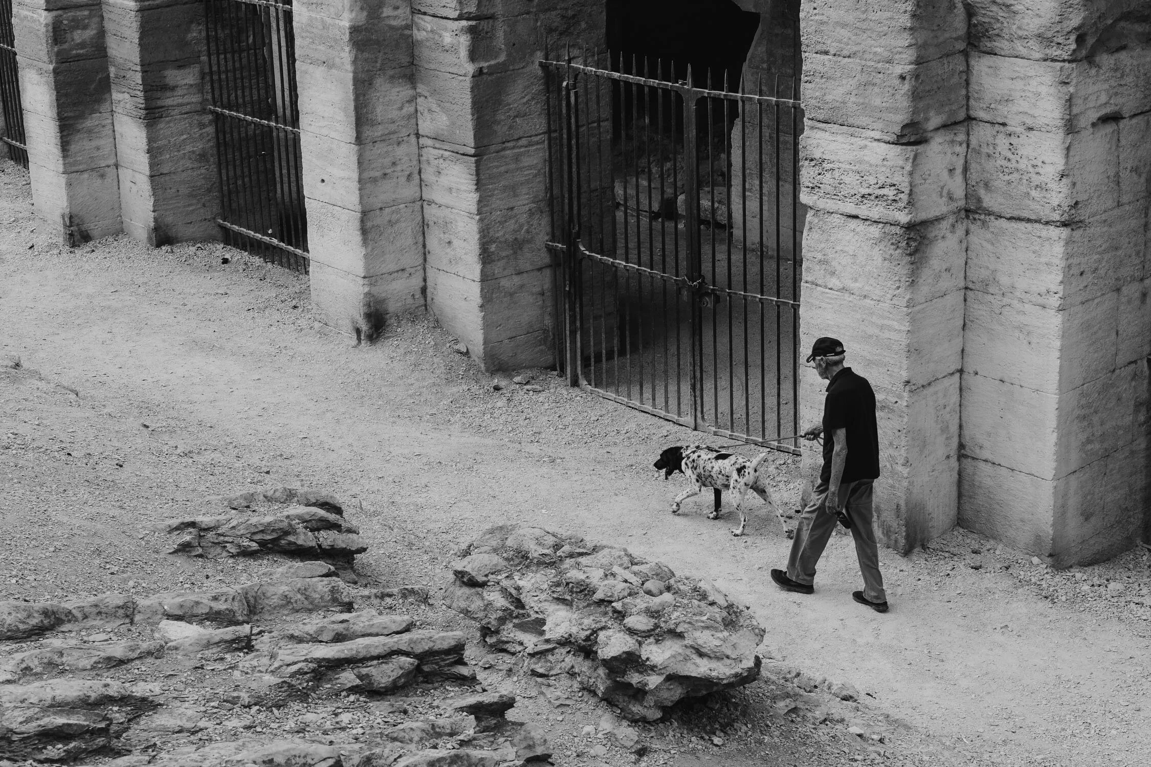 A man in a cap walks his Dalmatian dog past a large stone structure with a barred gate in black and white.