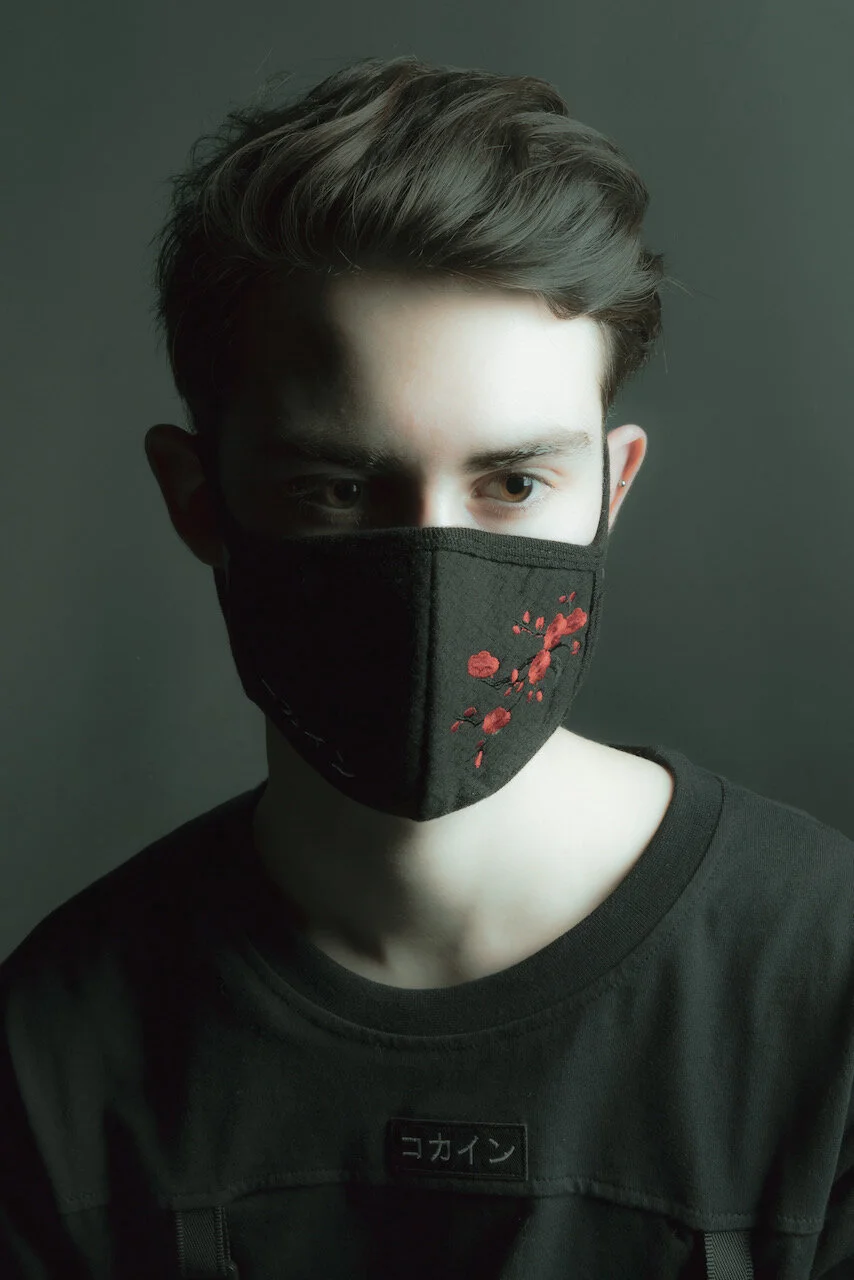A young man with styled dark hair wearing a black face mask with embroidered red flowers, black t-shirt, and a dark background.