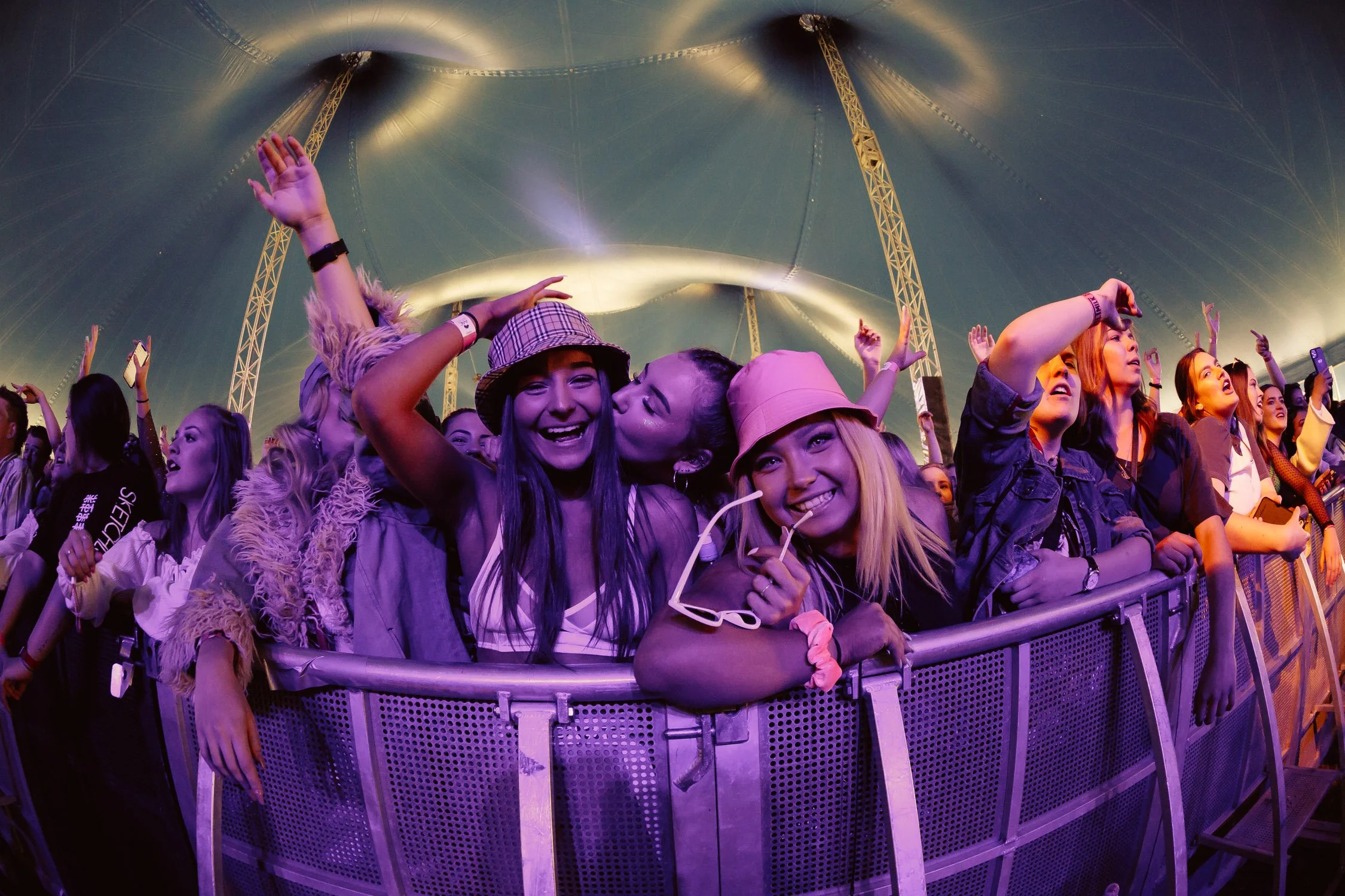Audience standing at a concert or festival inside a large tent, enjoying live music with some people dancing and smiling, under colorful lighting.