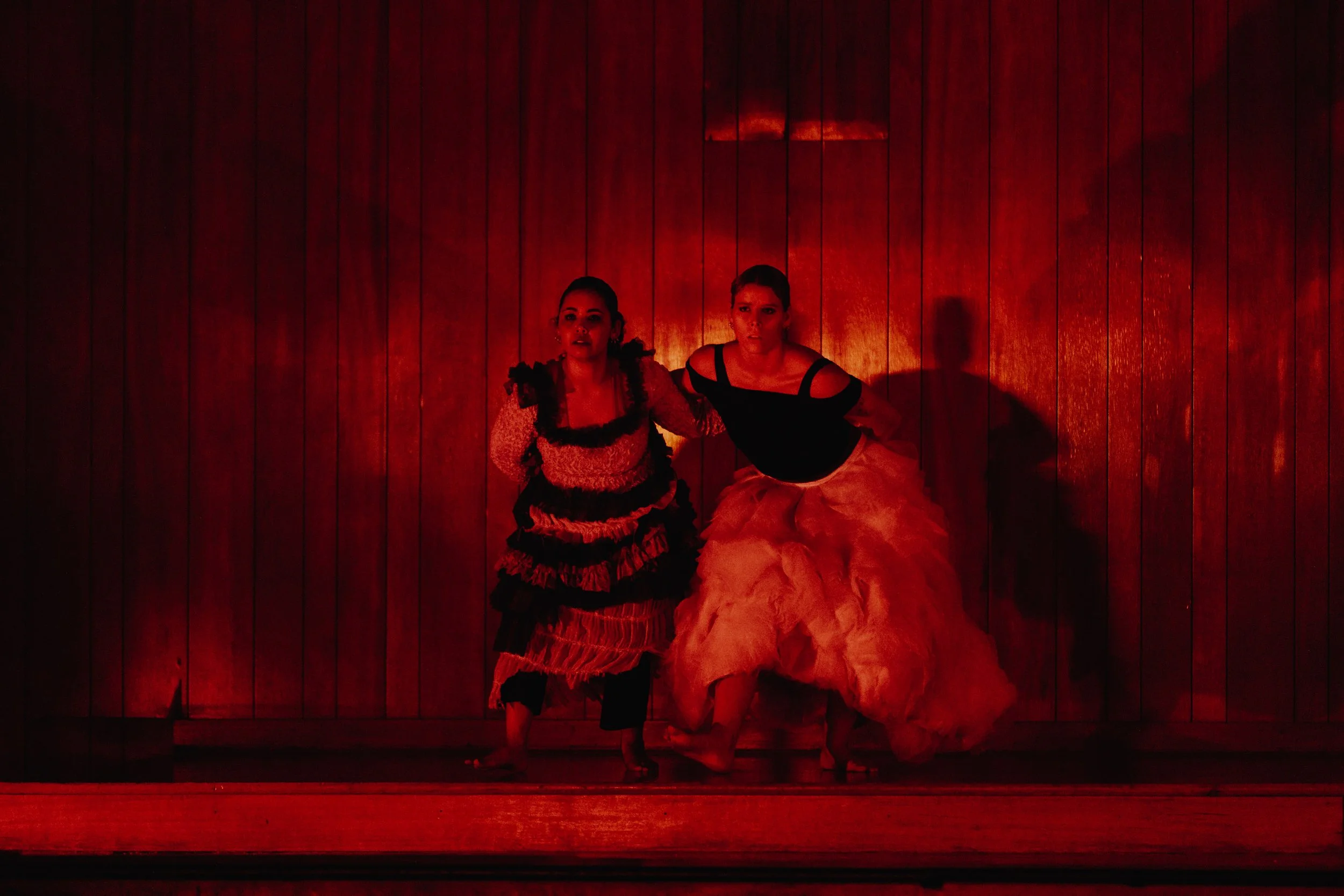 Two women in colorful costumes on stage with a wooden backdrop, illuminated by red lighting.
