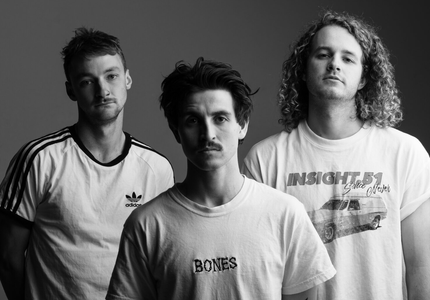 Black and white photo of three young men standing close together, facing forward. The man on the left wears a white Adidas t-shirt with black stripes, the man in the middle has a casual t-shirt with the word "BONES" on it, and the man on the right we