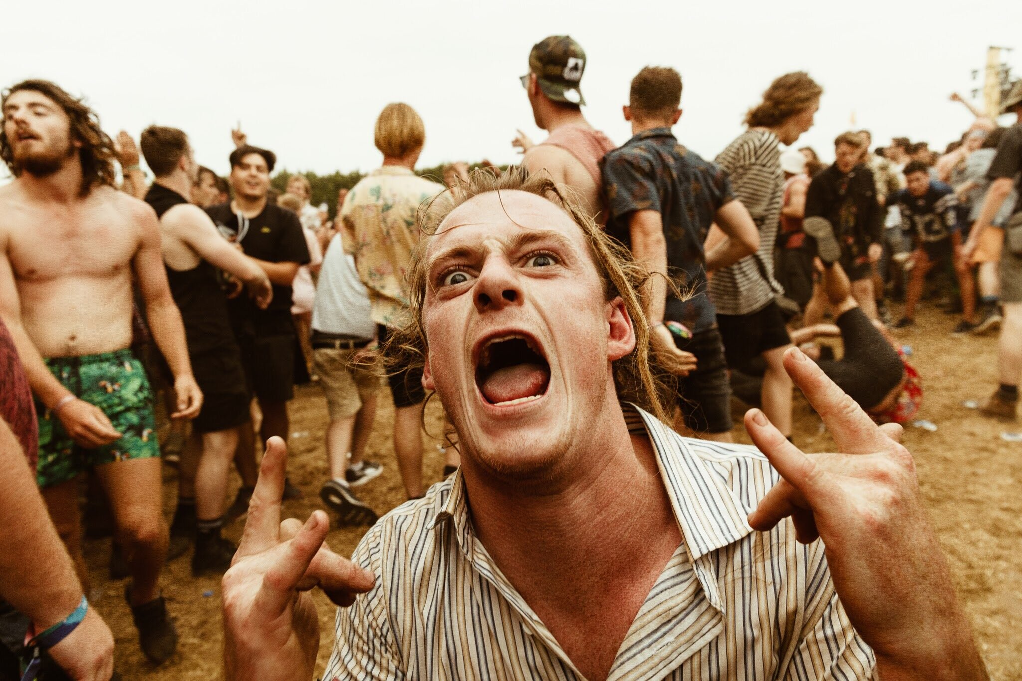 A man with long, light brown hair shouting with an intense expression at an outdoor event with many people in the background.