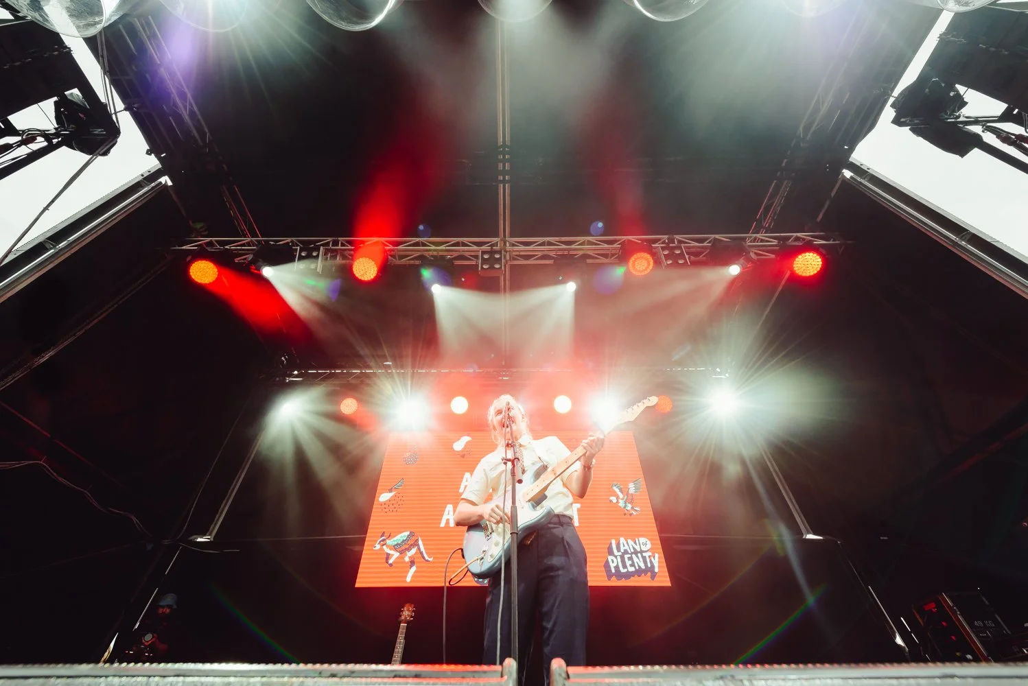 A musician playing an electric guitar on stage during a concert with bright red and white stage lighting and a large digital screen behind displaying graphics and text.