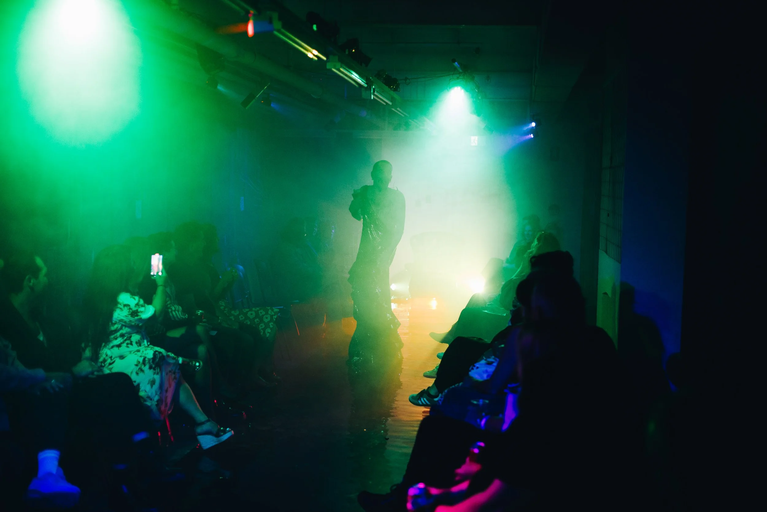 A performer standing on a stage illuminated by colorful green and yellow lights, with an audience seated and some taking photos in a dimly lit room.