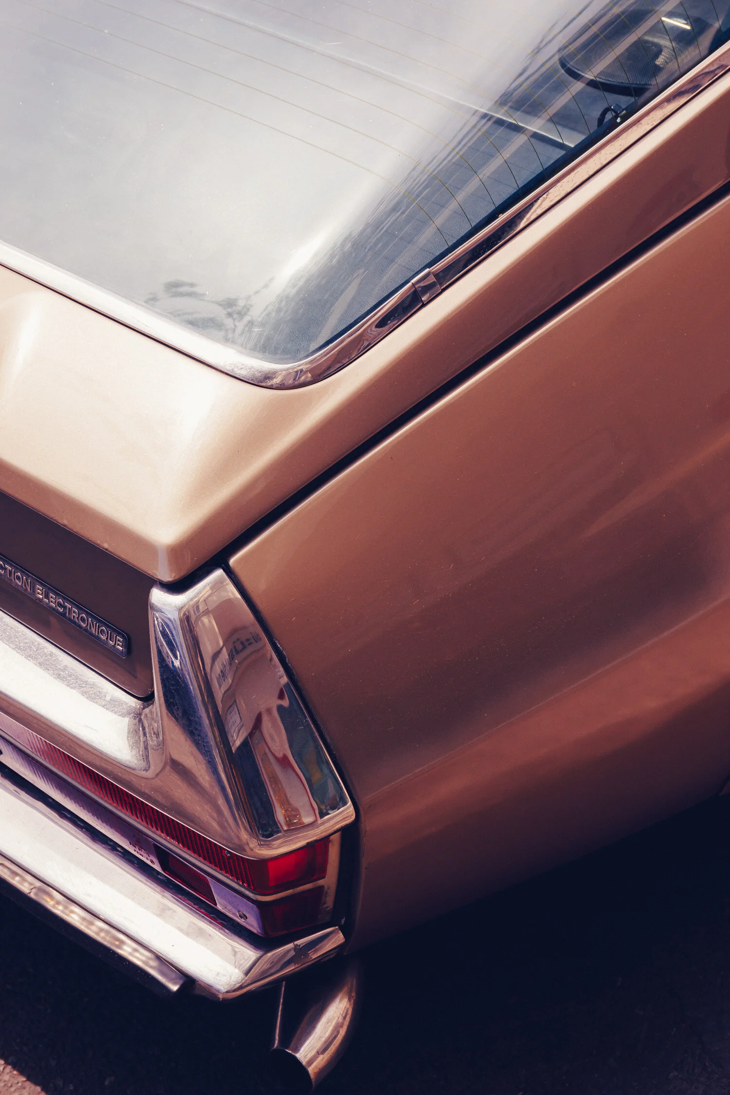 Close-up of the rear corner of a vintage gold-colored car, showing part of the trunk, tail light, and exhaust pipe.