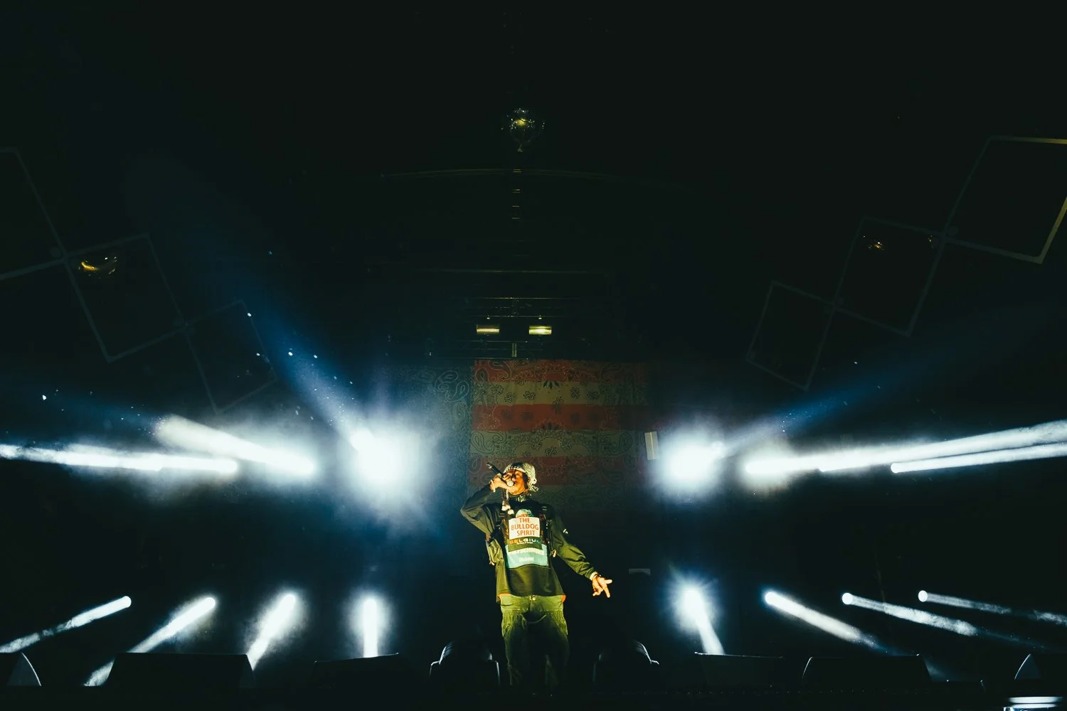 A performer on stage with bright white lights and fog, holding a microphone and wearing a dark jacket and a shirt with text, inside a dark concert venue.