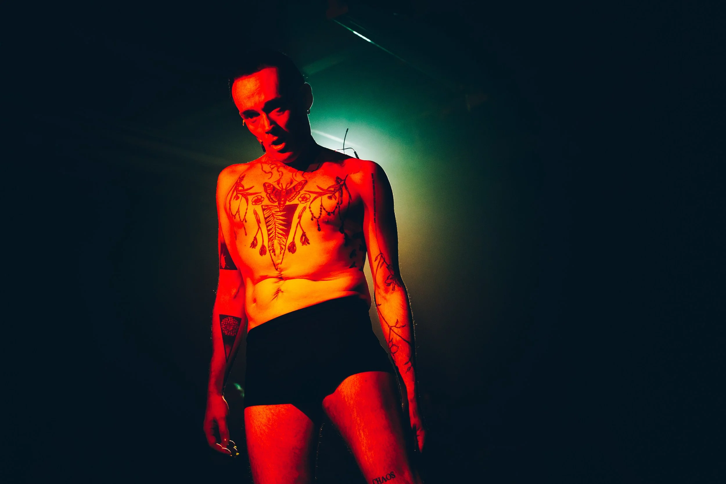 A shirtless person with numerous tattoos, wearing black shorts, illuminated by red and green lighting.