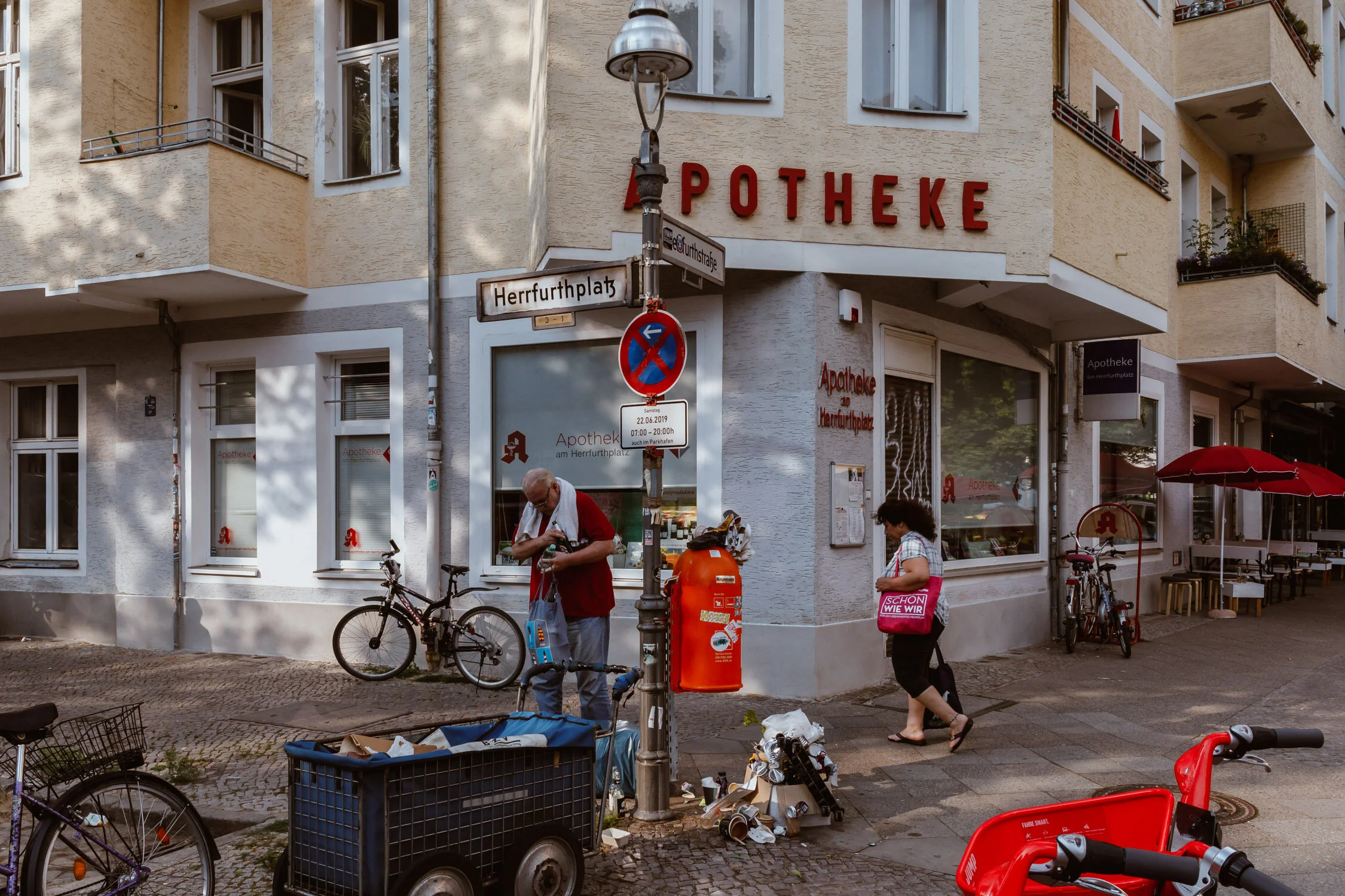 Street scene in front of a pharmacy with a red sign reading 'Apotheke'. An elderly man and a woman are on the sidewalk, with bicycles and a shopping cart nearby, and a street sign reading 'Herrfurthplatz'.