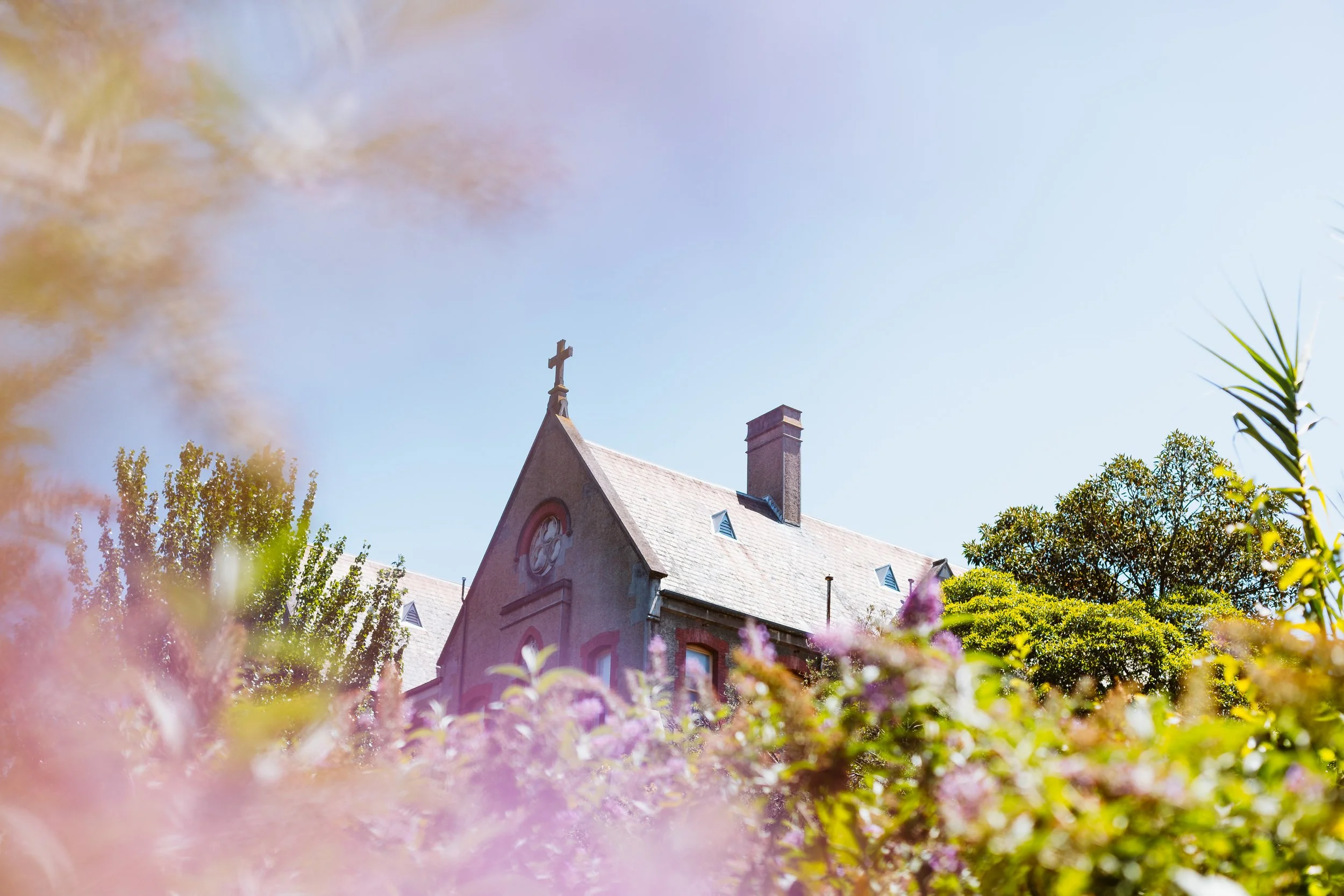 A church building with a cross on the peak of the roof, surrounded by trees and flowers under a clear blue sky.