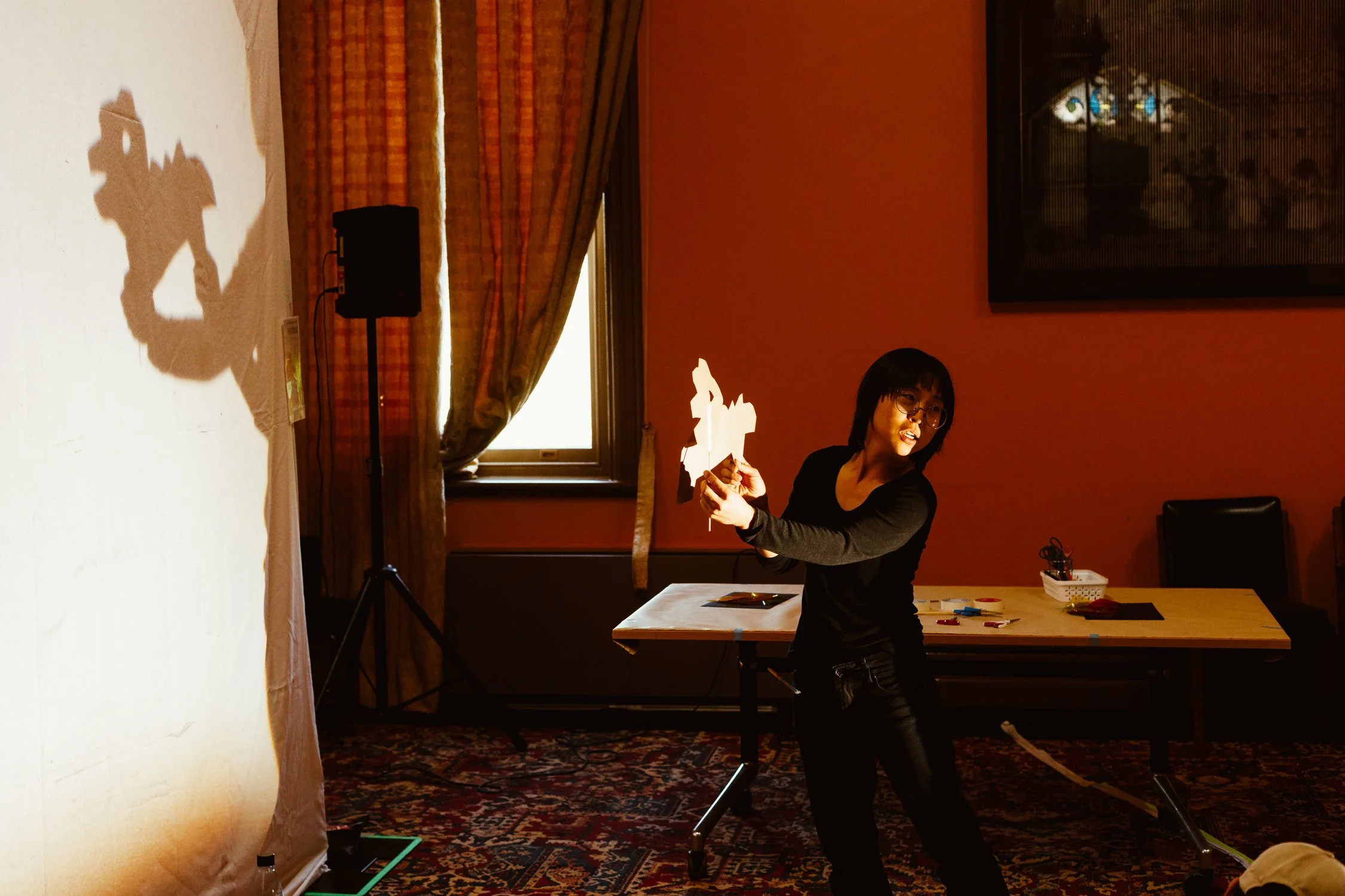 Woman with glasses and black shirt performing shadow puppet show with paper figures on an indoor stage, shadow cast on a white screen to her left, room decorated with patterned carpet, curtains, window, and a framed painting.