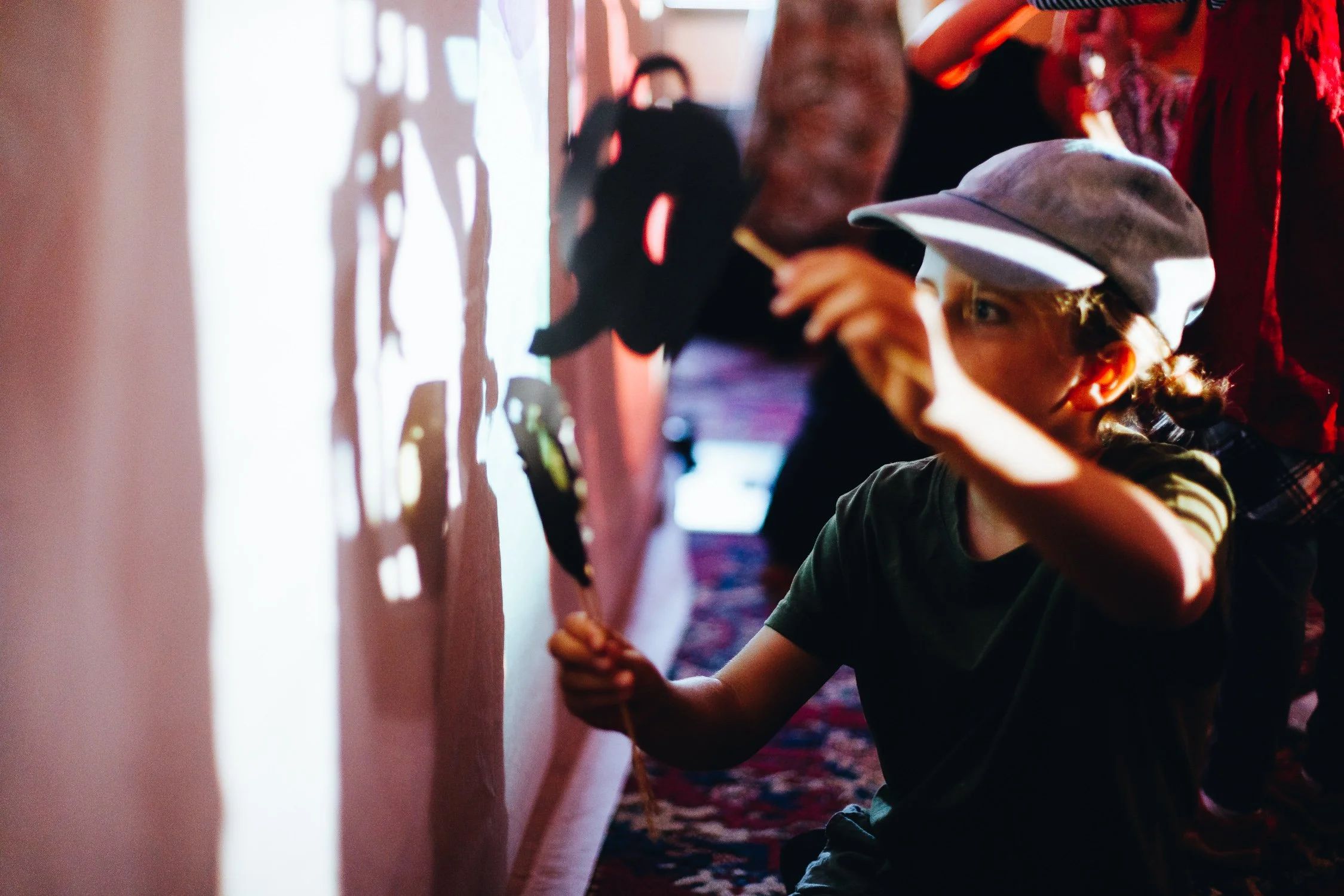A young girl in a gray cap and green shirt participates in a shadow puppet activity on a wall, with other children and adults nearby.