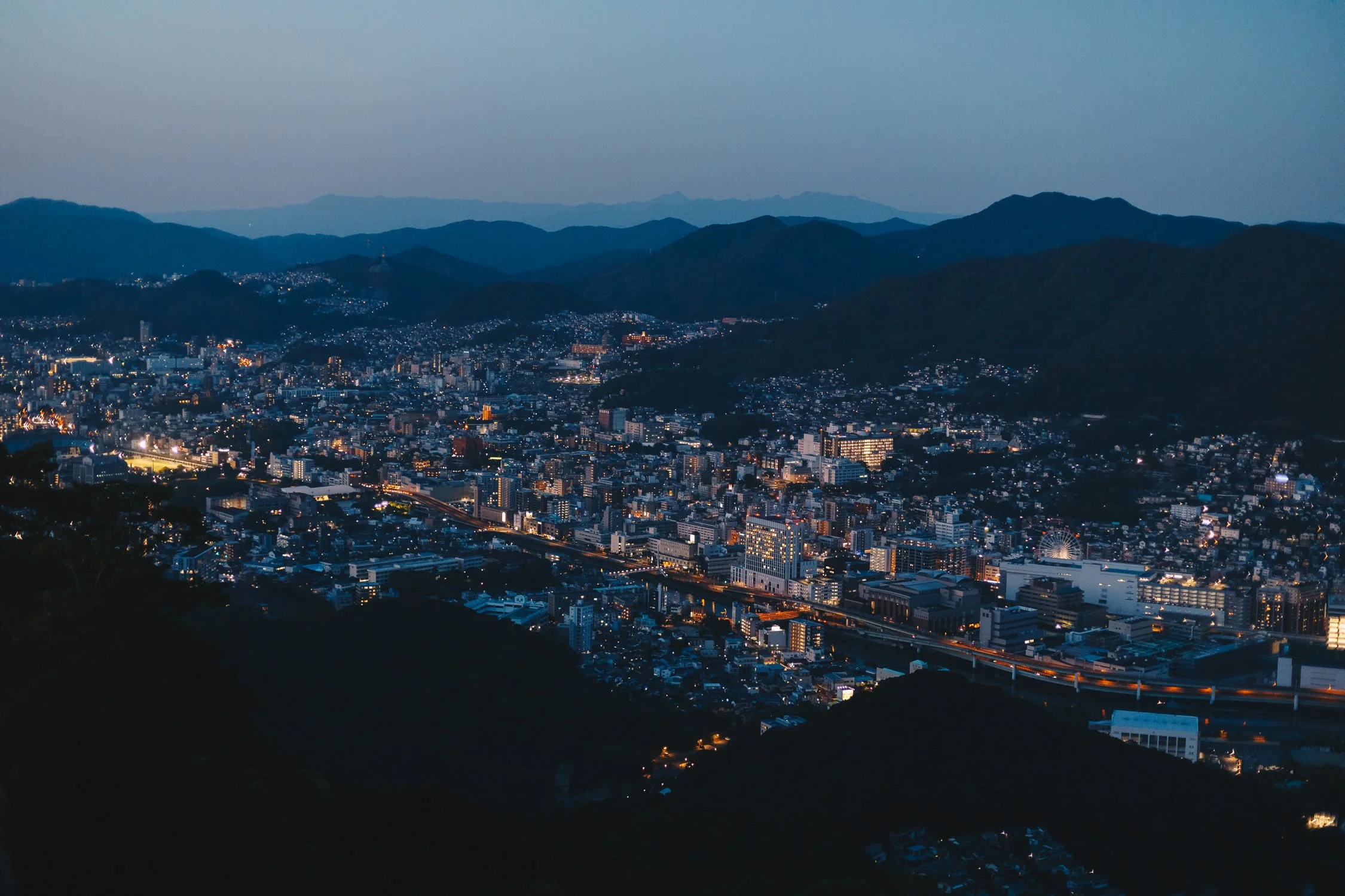 Cityscape at dusk with illuminated buildings and mountains in the background.