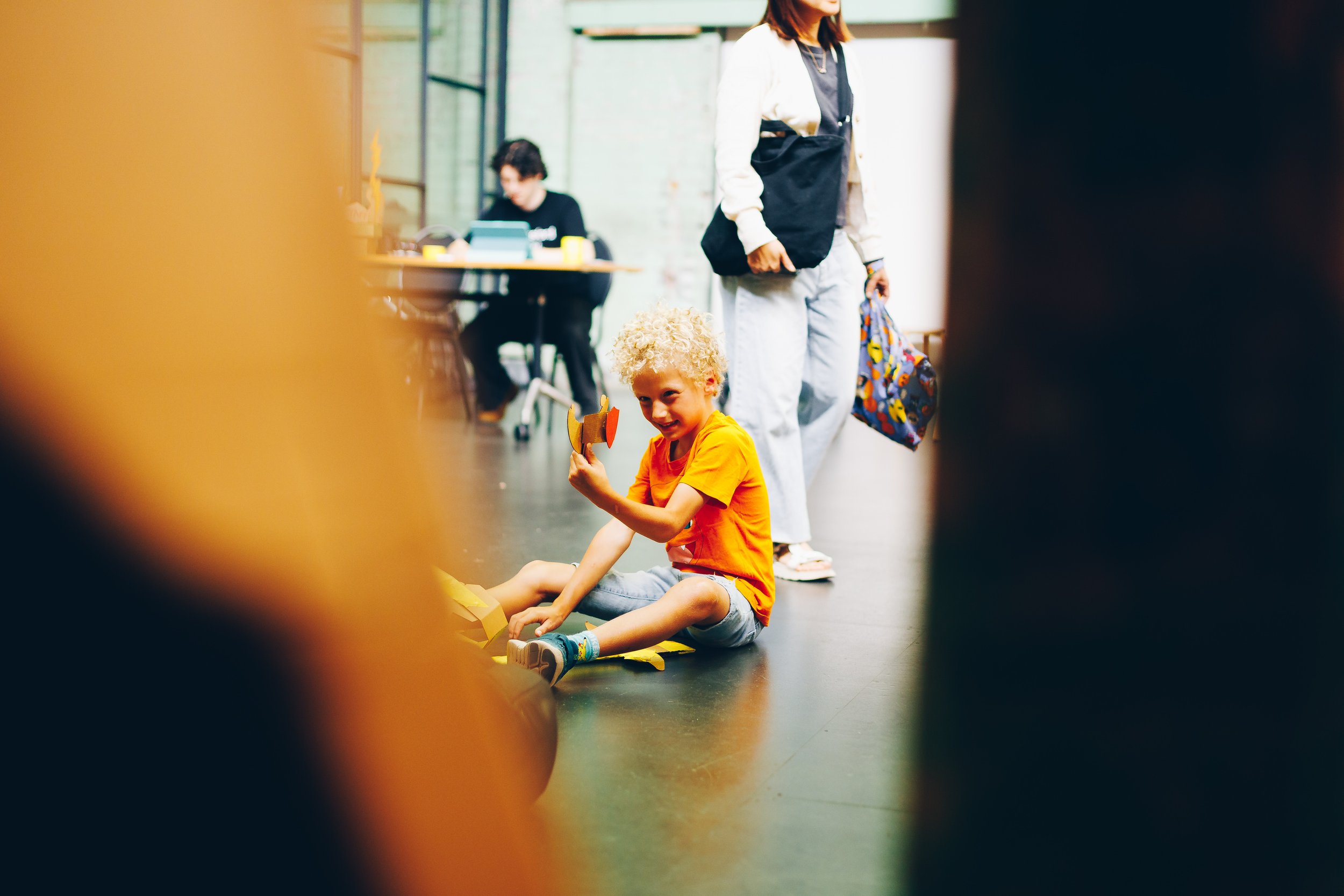 A young boy with curly blonde hair, wearing an orange T-shirt and gray shorts, sitting on the floor of an indoor space, smiling and holding a colorful toy. In the background, a woman in a white jacket and gray pants stands nearby, and a person workin