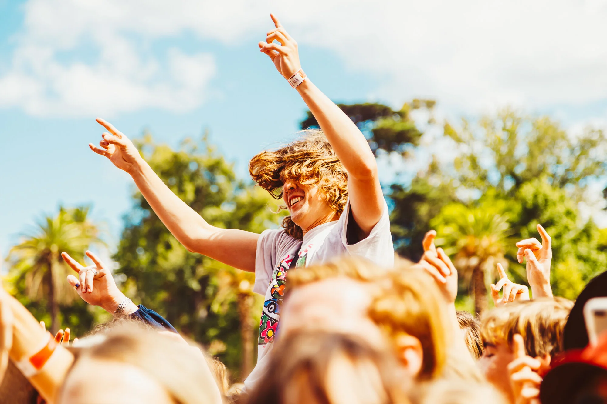 A woman with curly hair dancing at an outdoor event on a sunny day, surrounded by people, with trees and a blue sky in the background.