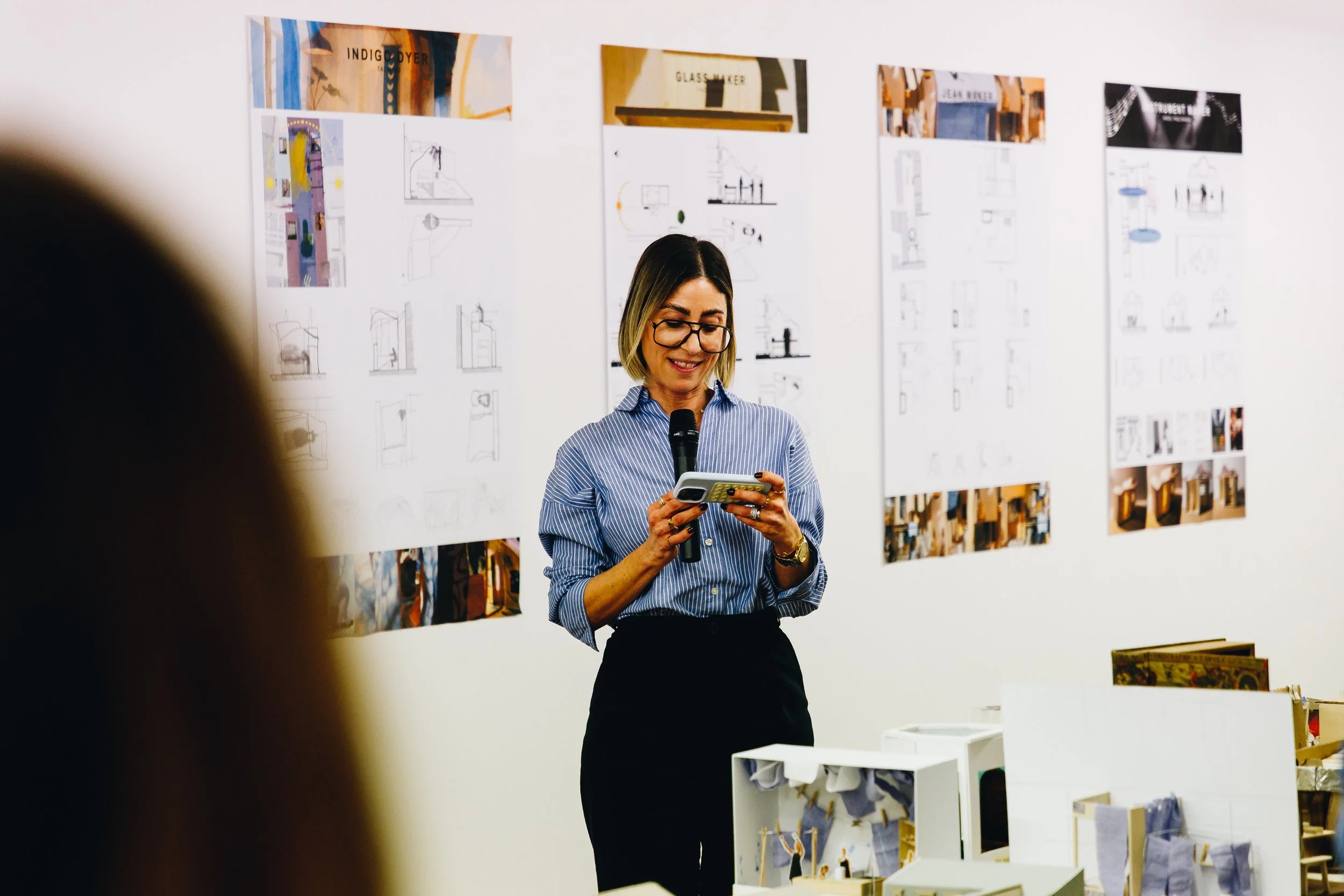 A woman with glasses, wearing a blue striped shirt, is standing in front of a white wall with various design boards and sketches. She is smiling and holding a microphone in one hand and a smartphone in the other.