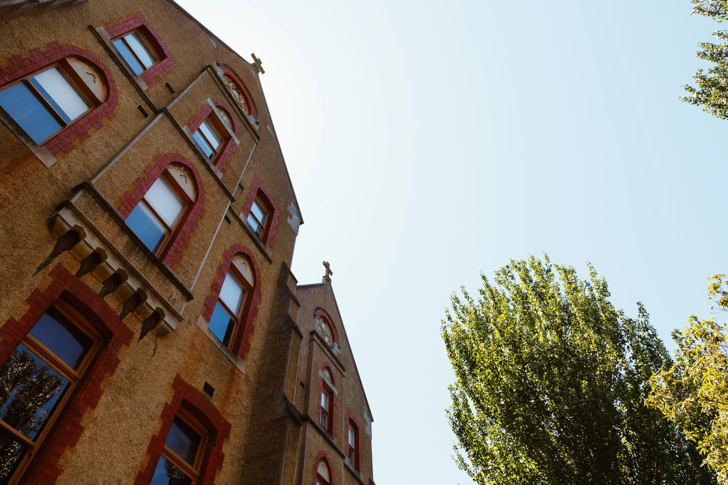 A tall historic building with brick and stucco exterior, arched windows with decorative brick trim, and small crosses on the roof, surrounded by trees against a clear blue sky.
