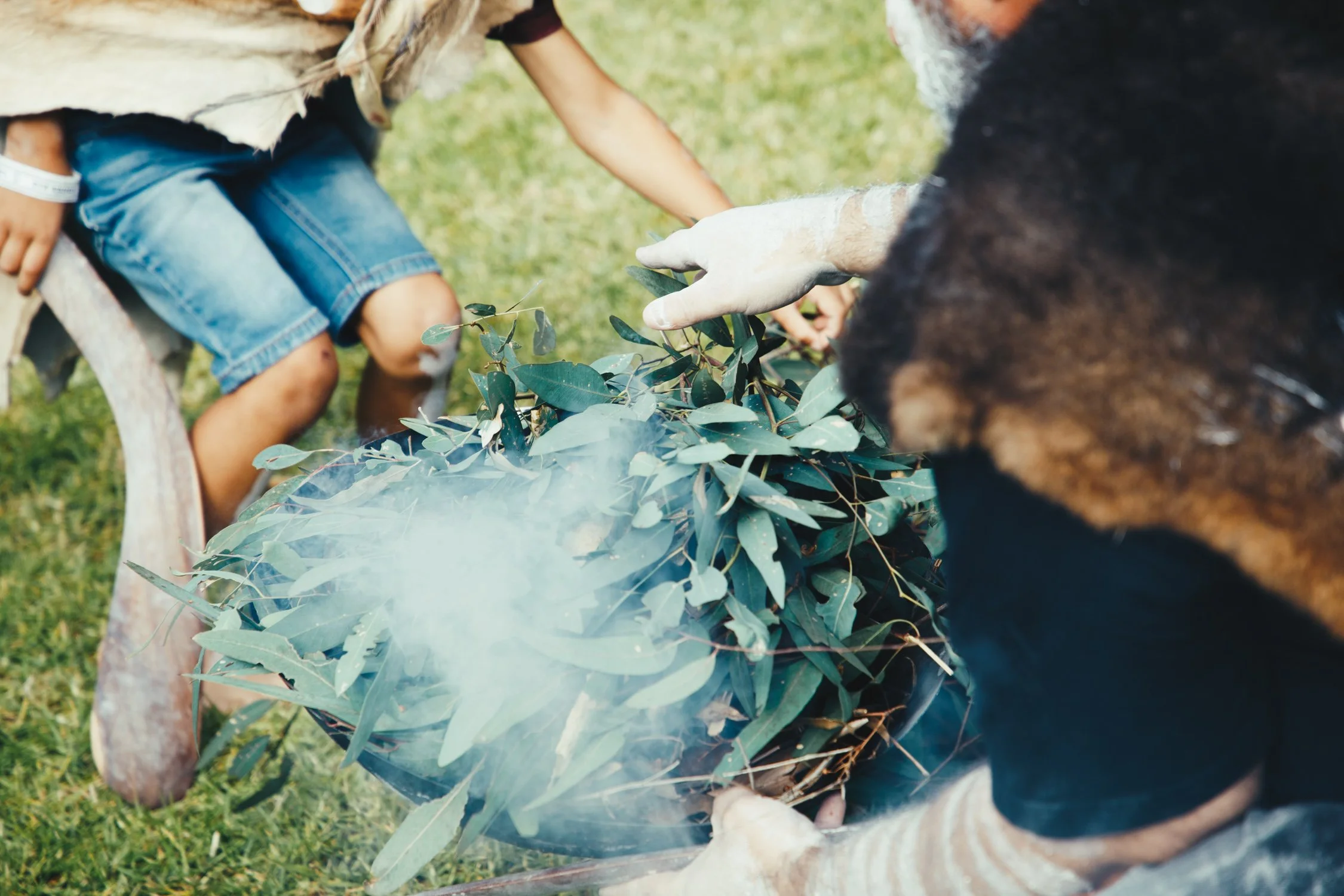 Two people and a child handling a bundle of green leaves outdoors with smoke rising from it.