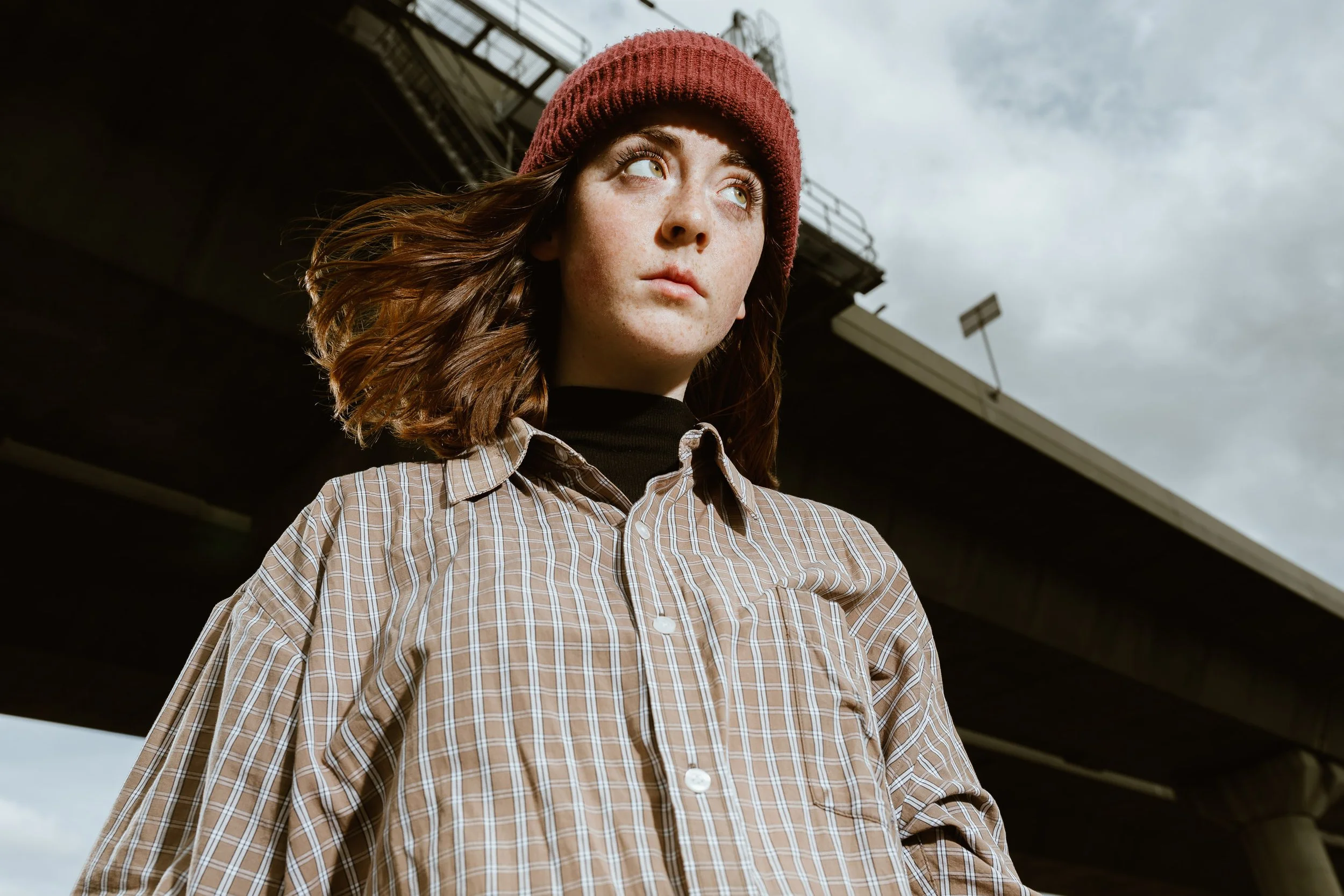 Young woman with shoulder-length brown hair, wearing a red knit beanie, a black turtleneck, and a beige plaid shirt, standing outdoors beneath a bridge with overcast skies in the background.