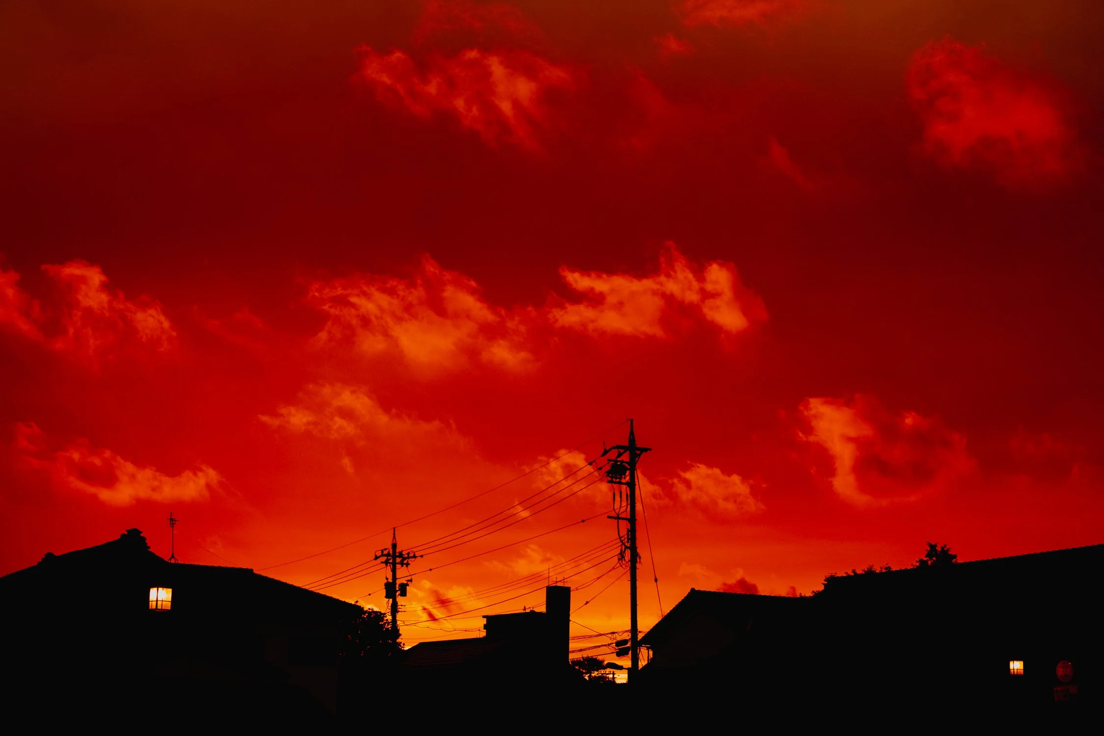 Silhouettes of houses and utility poles against a vivid red and orange sunset sky with scattered clouds.