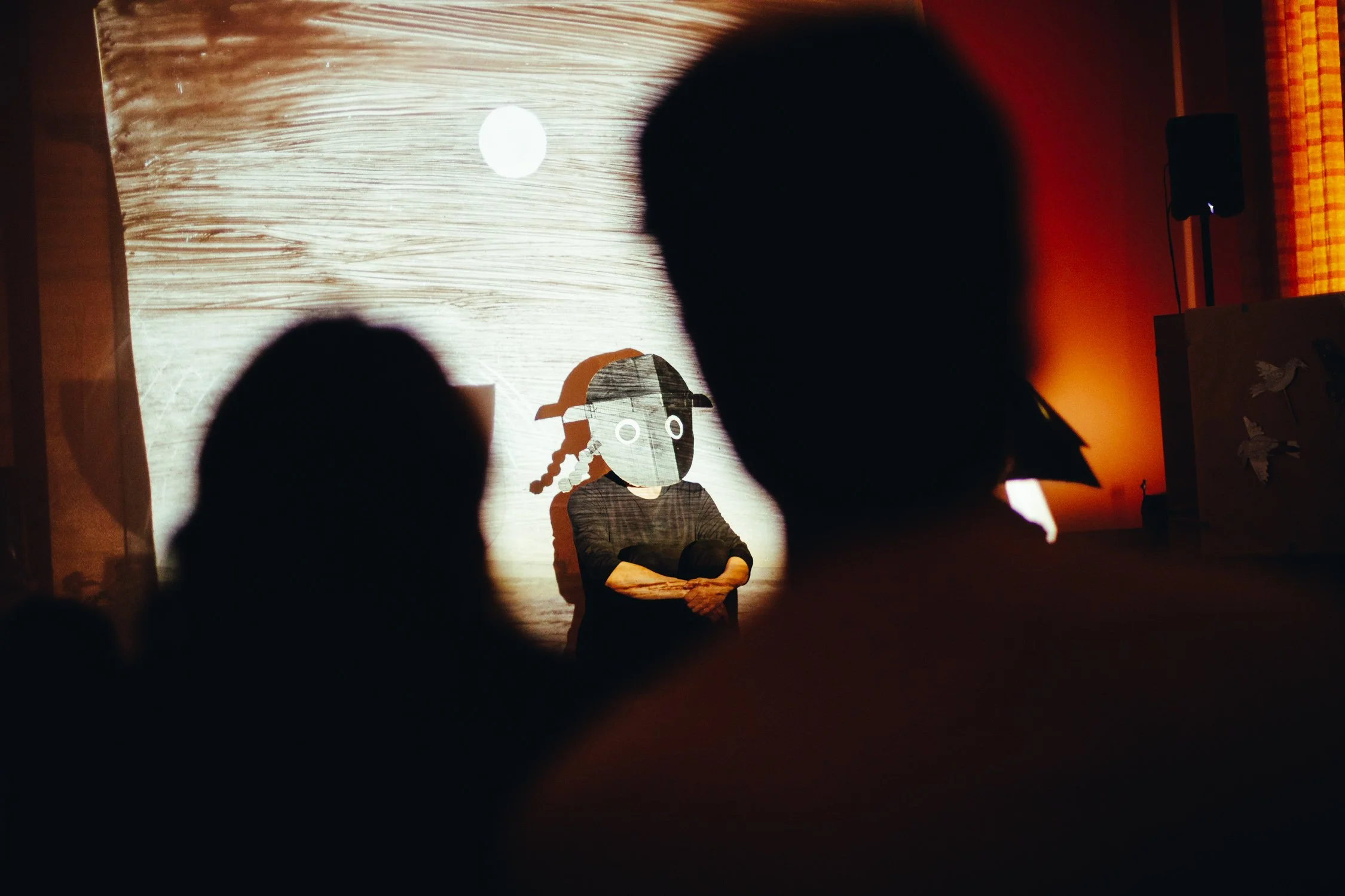 Silhouette of children watching shadow puppet show with a backdrop of a person wearing a puppet mask on stage.