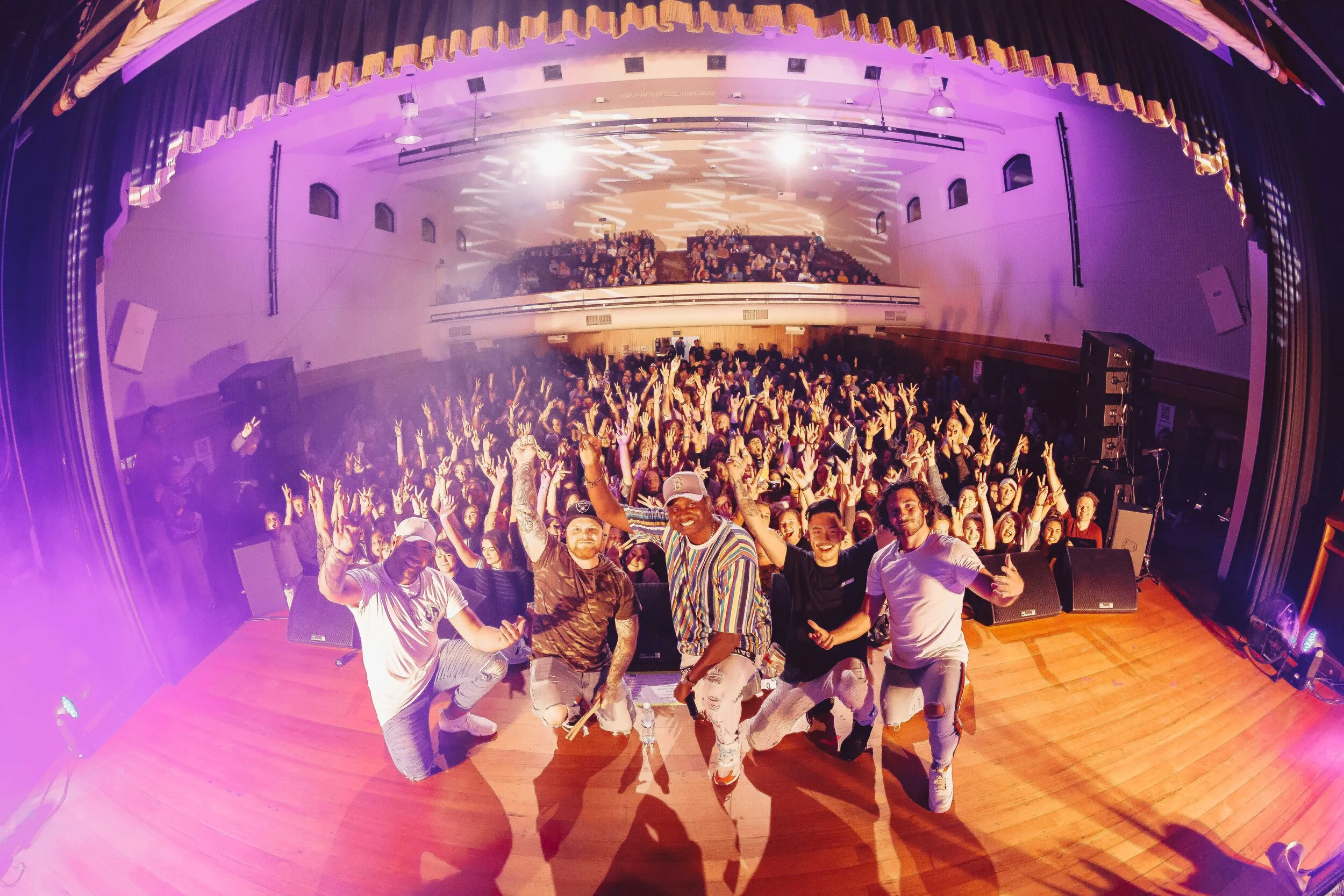 A group of performers taking a selfie on stage in front of a large audience in a concert hall with purple lighting and curtains.