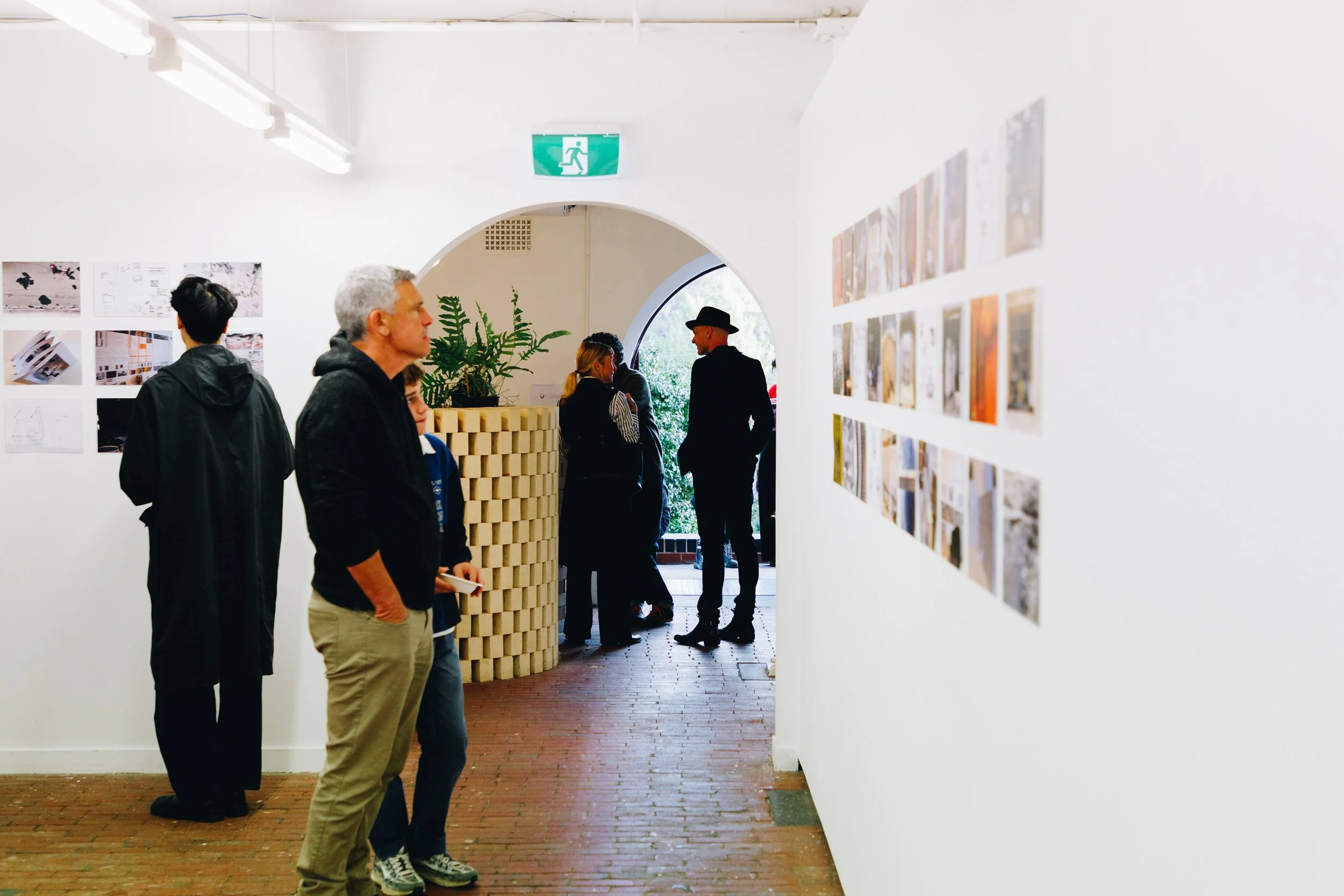 People viewing artwork and photographs in an art gallery, with white walls and bright overhead lighting, and an exit sign visible in the background.