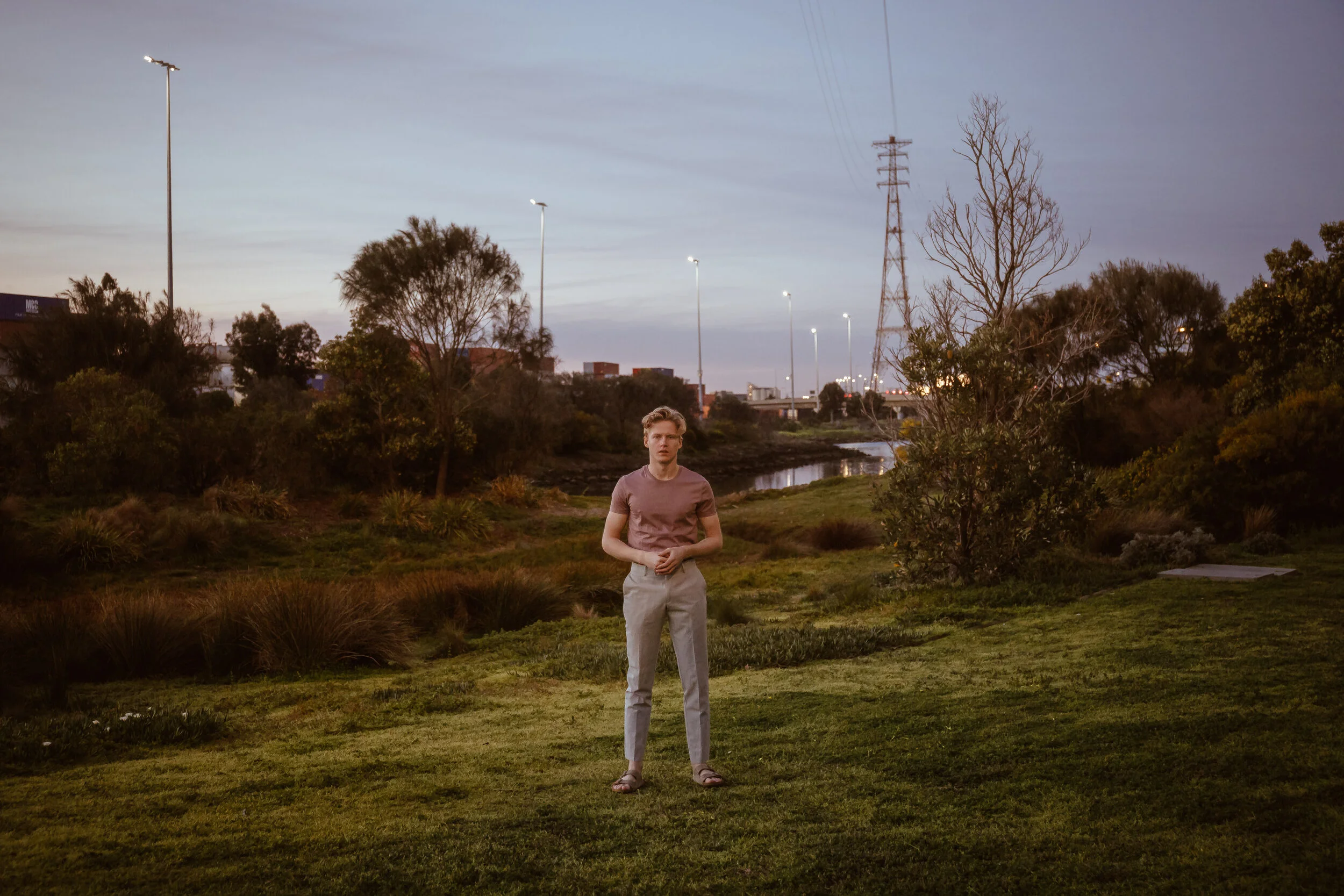 A young man standing in a grassy park during dusk with trees, a small river, and a cityscape with tall streetlights and power lines in the background.
