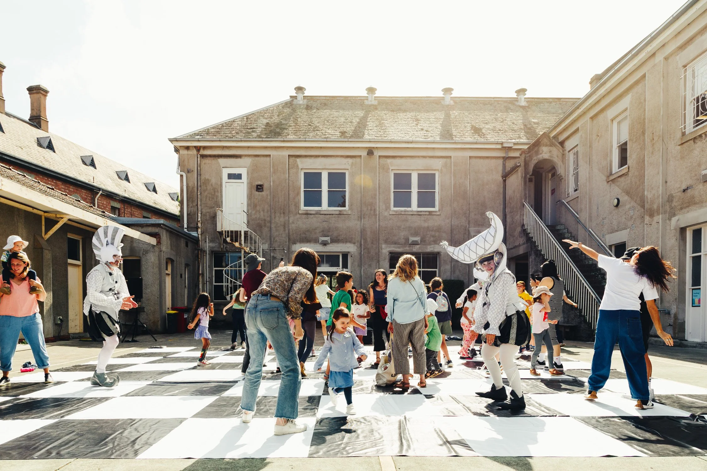 People playing chess in an outdoor courtyard with buildings in the background and sunlight overhead.