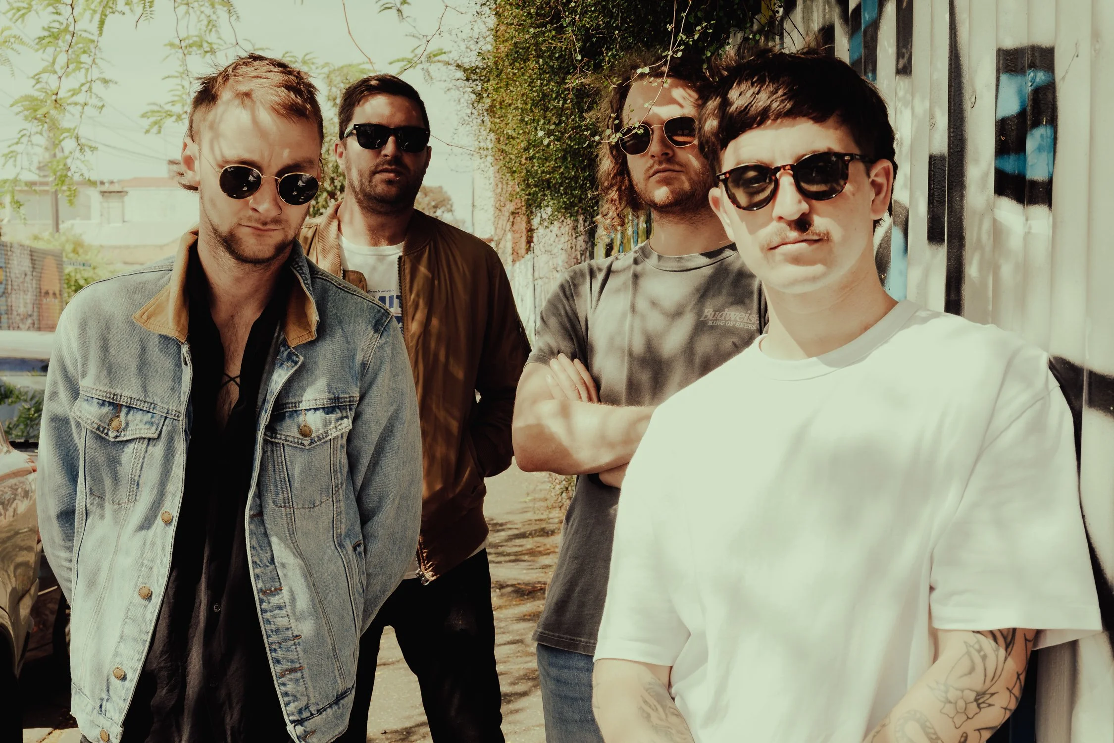 Four young men standing outdoors, wearing sunglasses, with graffiti-covered walls and trees in the background.