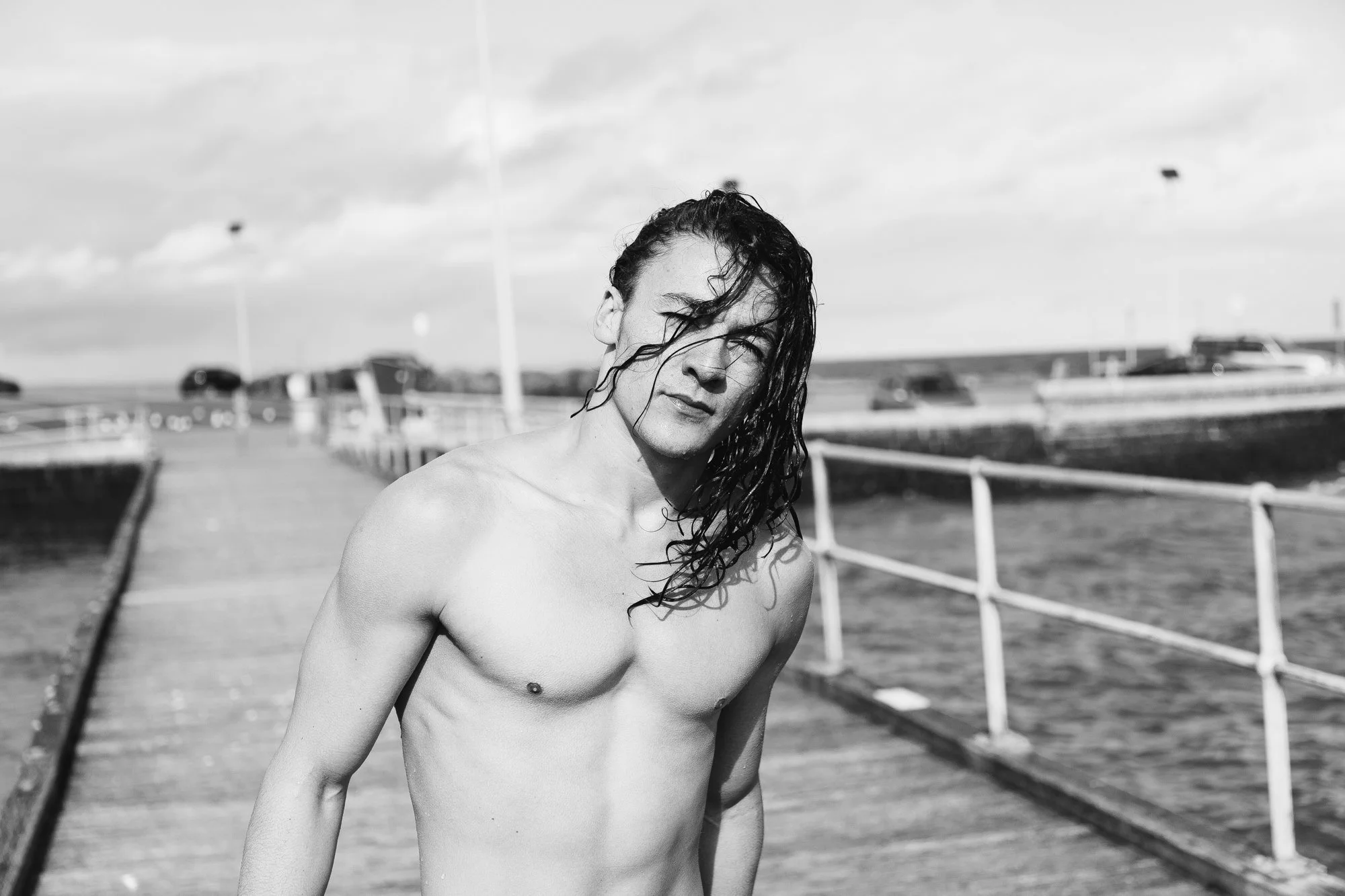 A shirtless young man with wet hair standing on a pier near water, with boats and a cloudy sky in the background.
