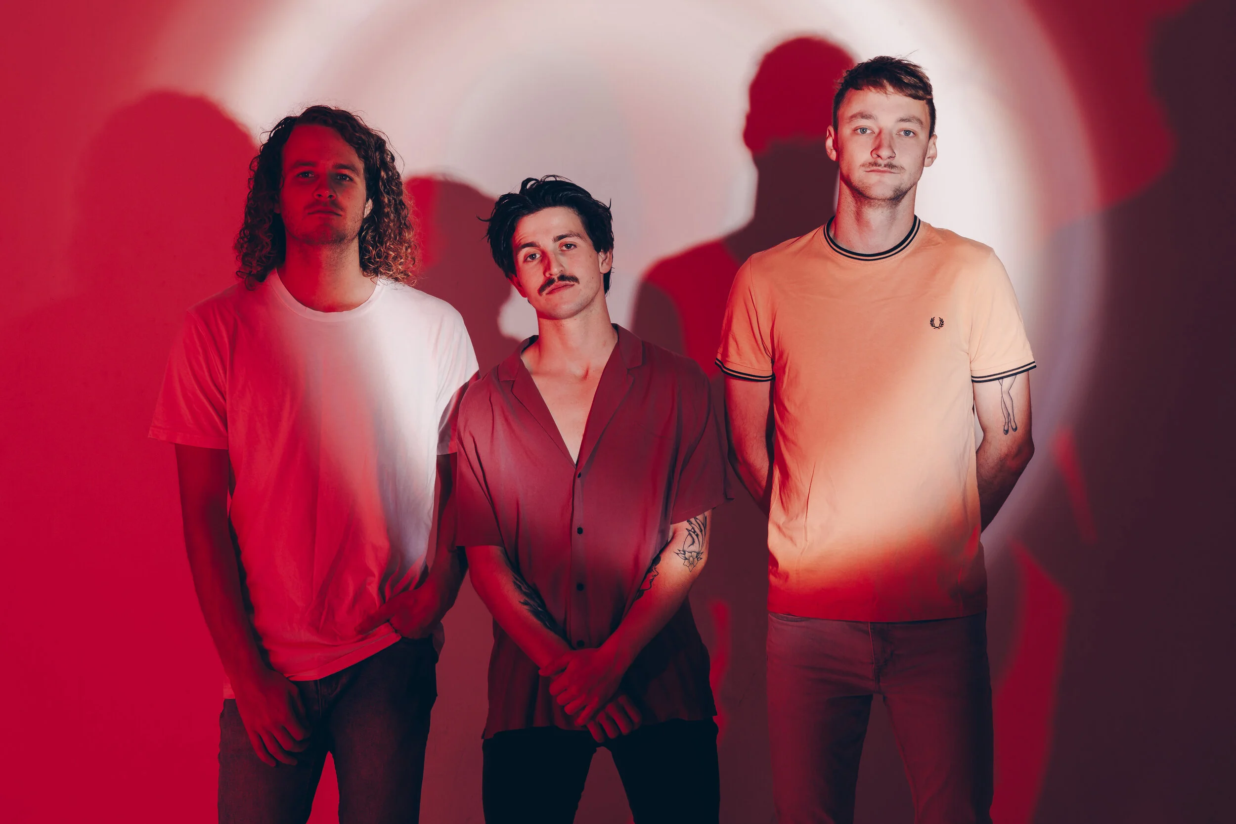 Three young men standing side by side against a wall with red and white lighting.