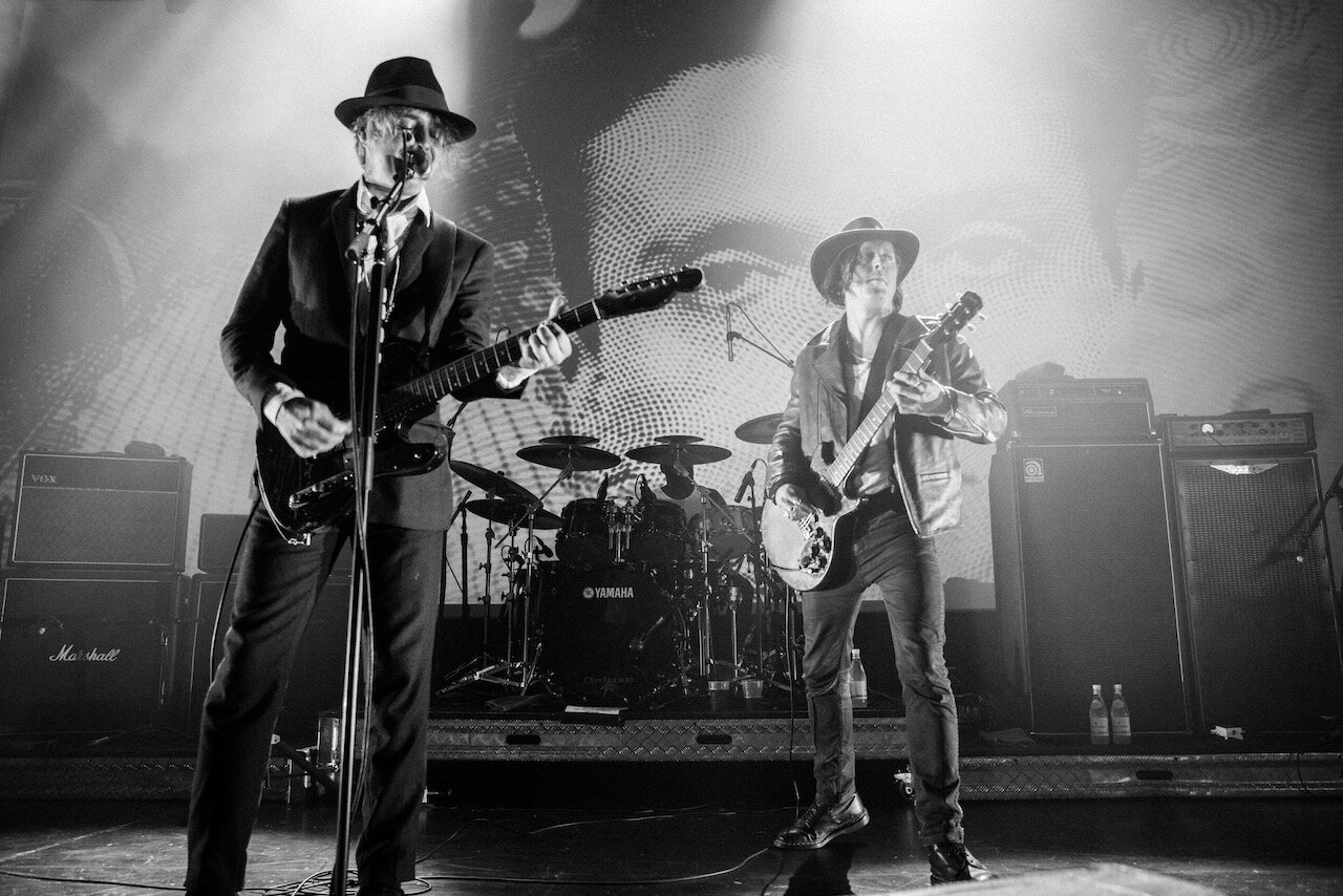 Two musicians perform on stage, each playing an electric guitar, with a drummer in the background. One musician is singing into a microphone. The stage has amplifiers and effect pedals, and there is a large screen or backdrop behind them.