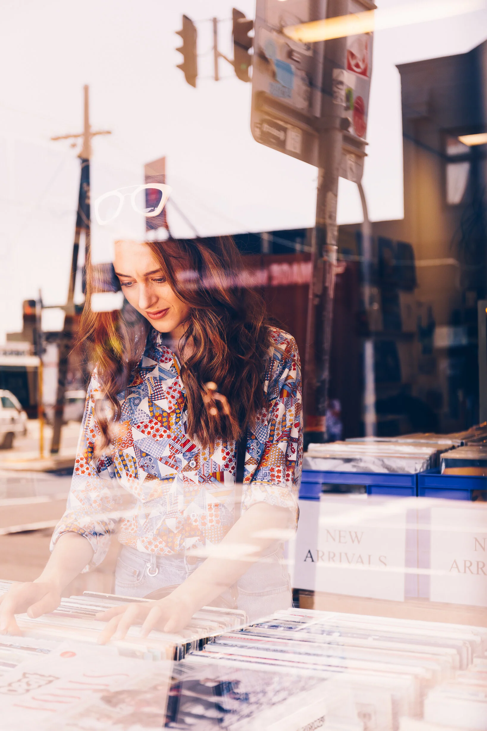 A young woman with long, wavy brown hair browsing magazines at a store window display, reflected with cars and street signs outside in an urban setting.