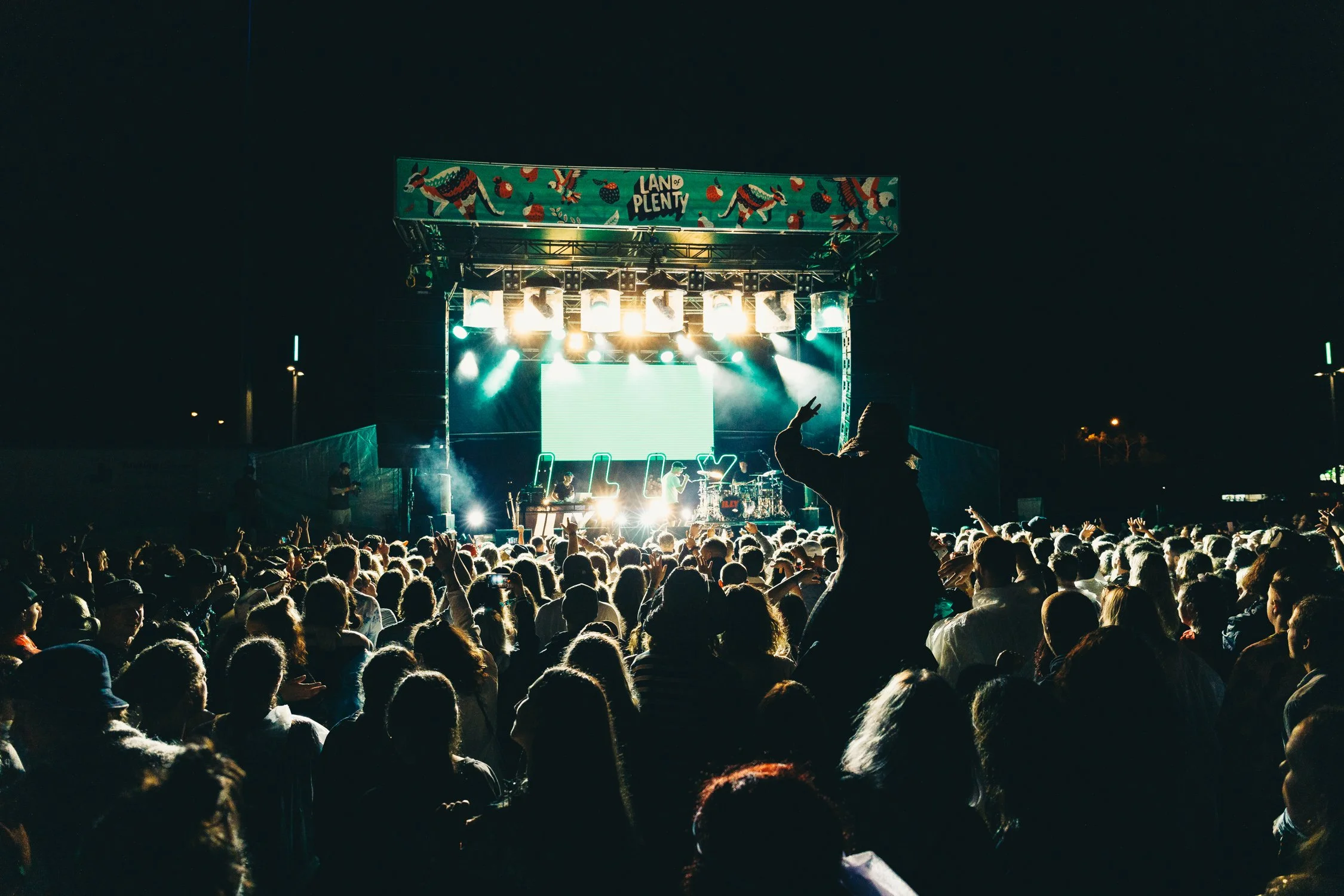 Crowd of people attending an outdoor concert at night with a brightly lit stage and a colorful banner that reads 'Land of Plenty' at the top.