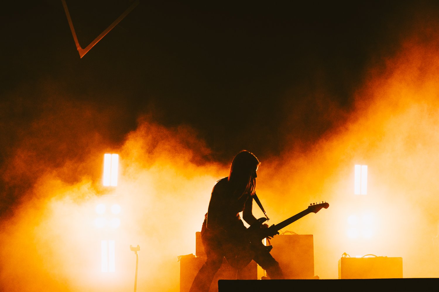A female guitarist performing on stage with bright orange lighting and smoke, silhouette visible against the illuminated background.