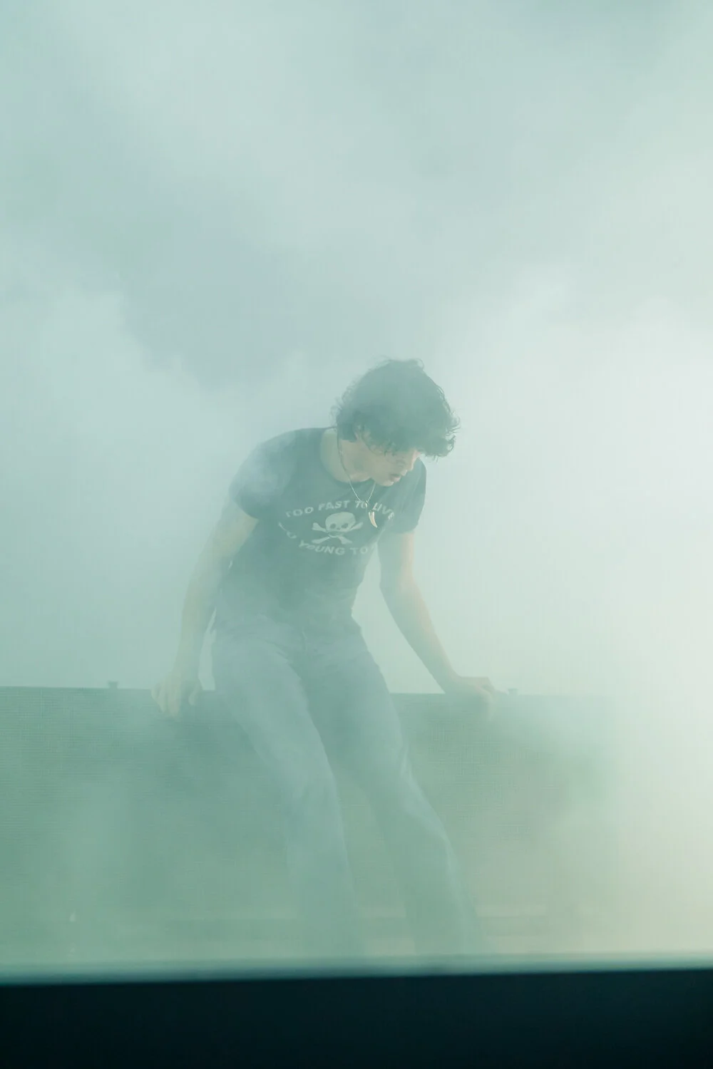 A person with curly dark hair crouches on a surface amidst dense smoke or fog, wearing a black t-shirt with a skull and crossbones graphic and text, with one hand on the surface and the other hanging down.