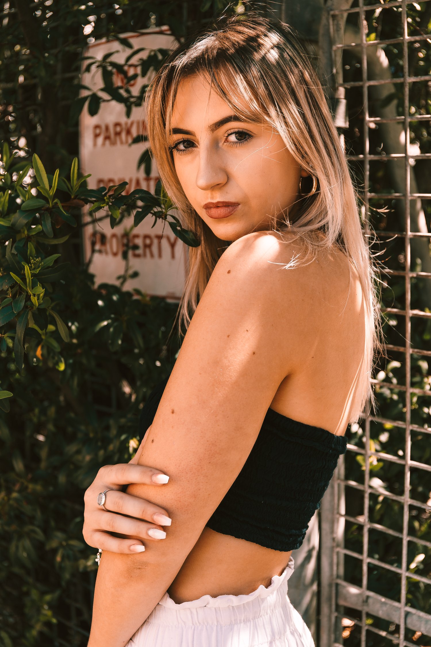 A young woman with blonde hair and tattoos on her arm is posing outdoors near a metal fence and green foliage, looking at the camera with a serious expression.