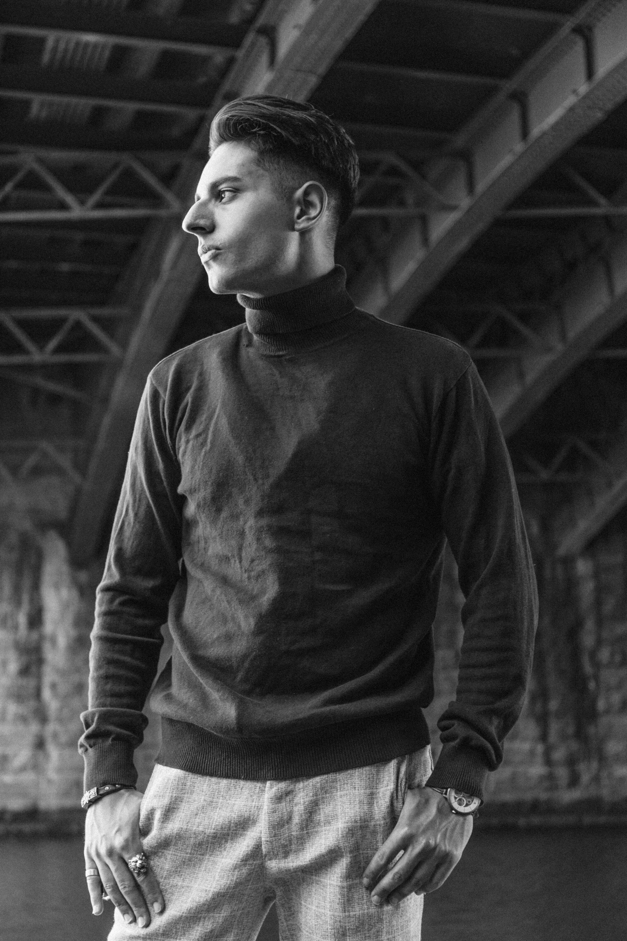 A young man in a black turtleneck sweater and light-colored pants standing under a bridge, looking to the side, in black and white.