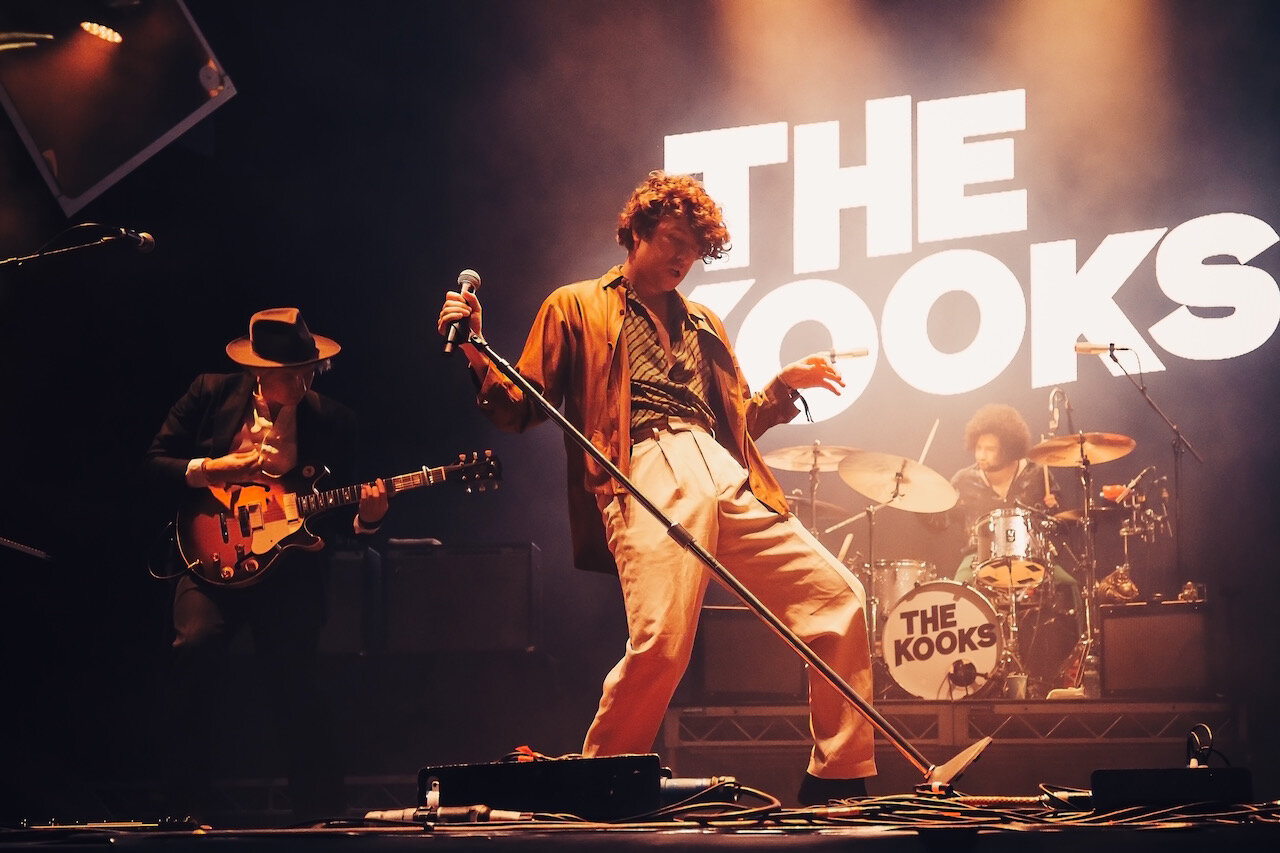 A band named The Kooks performing live on stage with a lead singer, guitarist, and drummer, with their name displayed in large lights behind them.