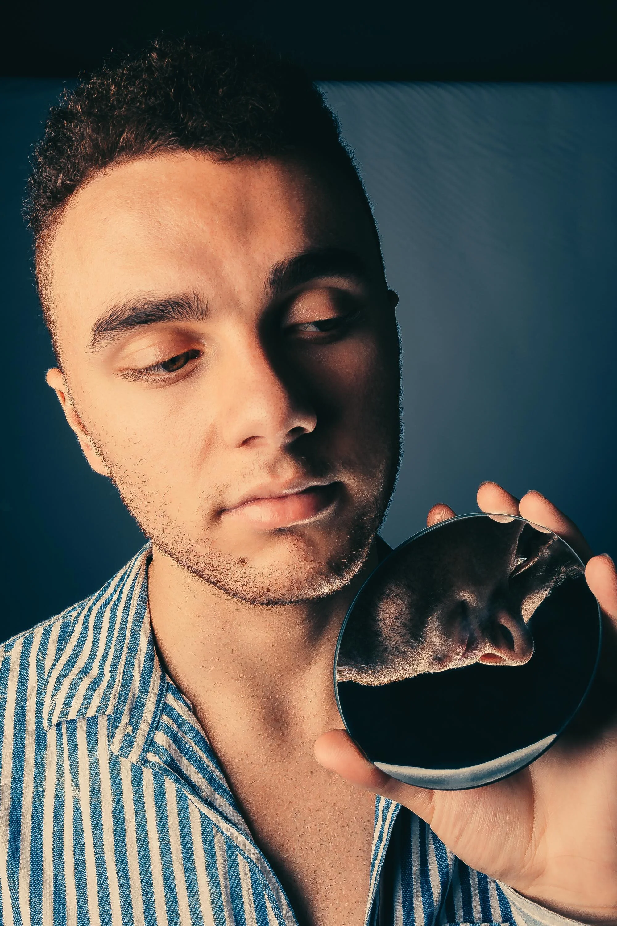 A young man with short, curly hair and light skin looks thoughtfully at a compact mirror he is holding in his hand. He wears a blue and white striped shirt and is posed against a plain background.