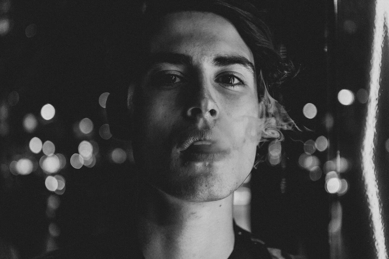 A close-up black and white photo of a young man with medium-length hair looking into the camera with a serious expression, with city lights blurred in the background.
