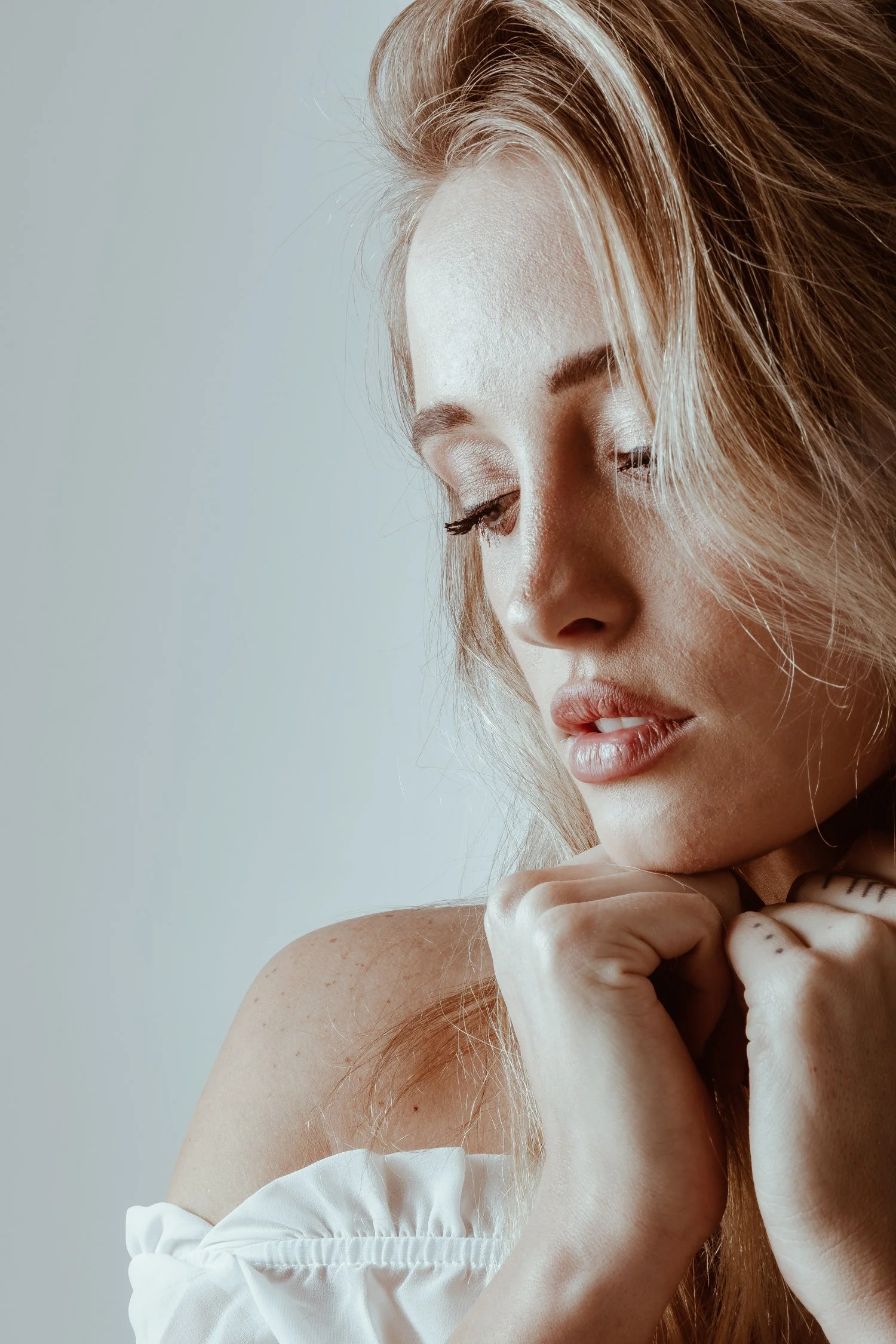 A close-up photo of a young woman with blonde hair, closed eyes, and lightly parted lips, resting her chin on her hand against a plain background.