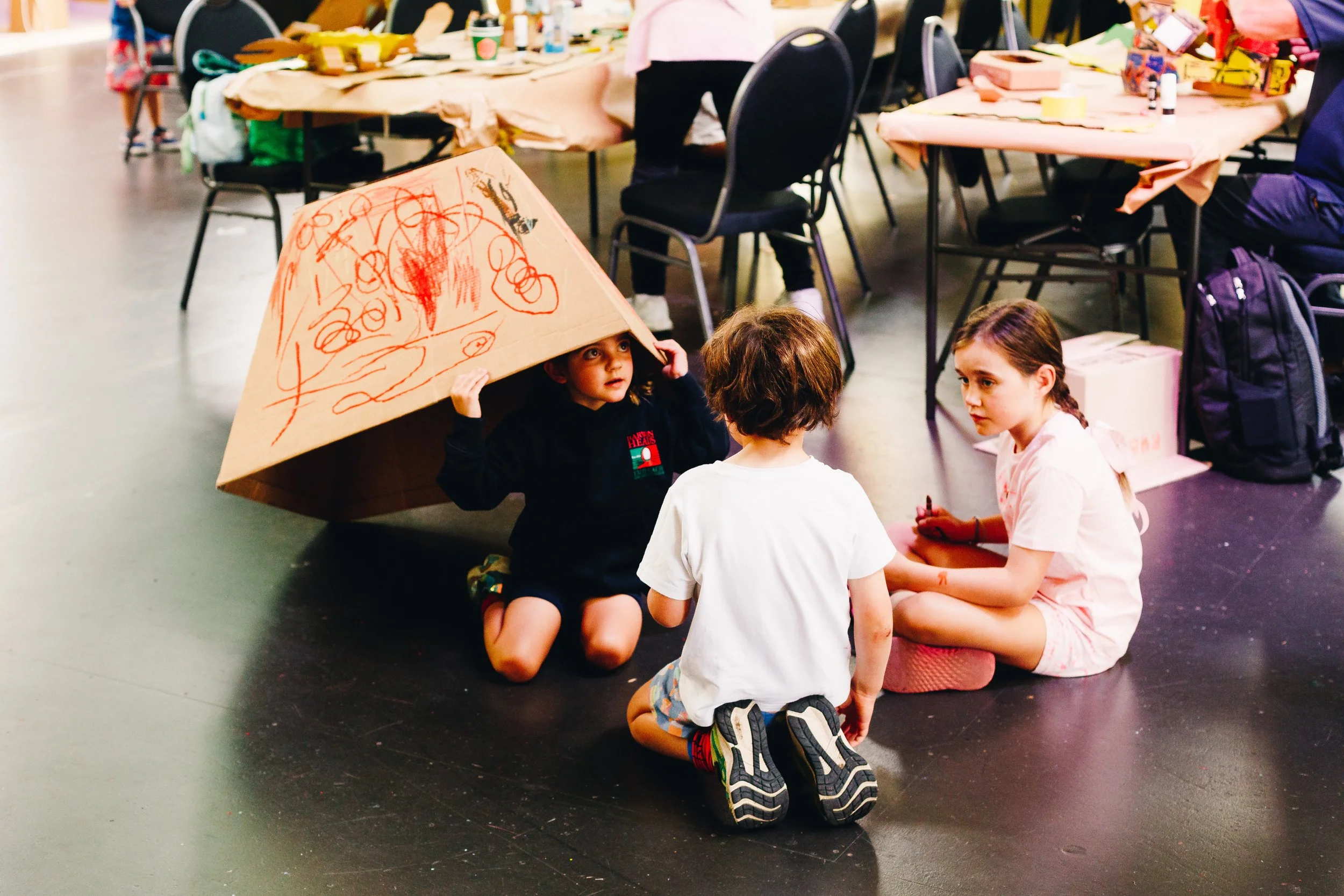 Three children sitting on the floor, engaging in conversation. One child is holding a large decorated cardboard box over their head as a hat. The room appears to be a gathering or event space with tables, chairs, and art supplies in the background.
