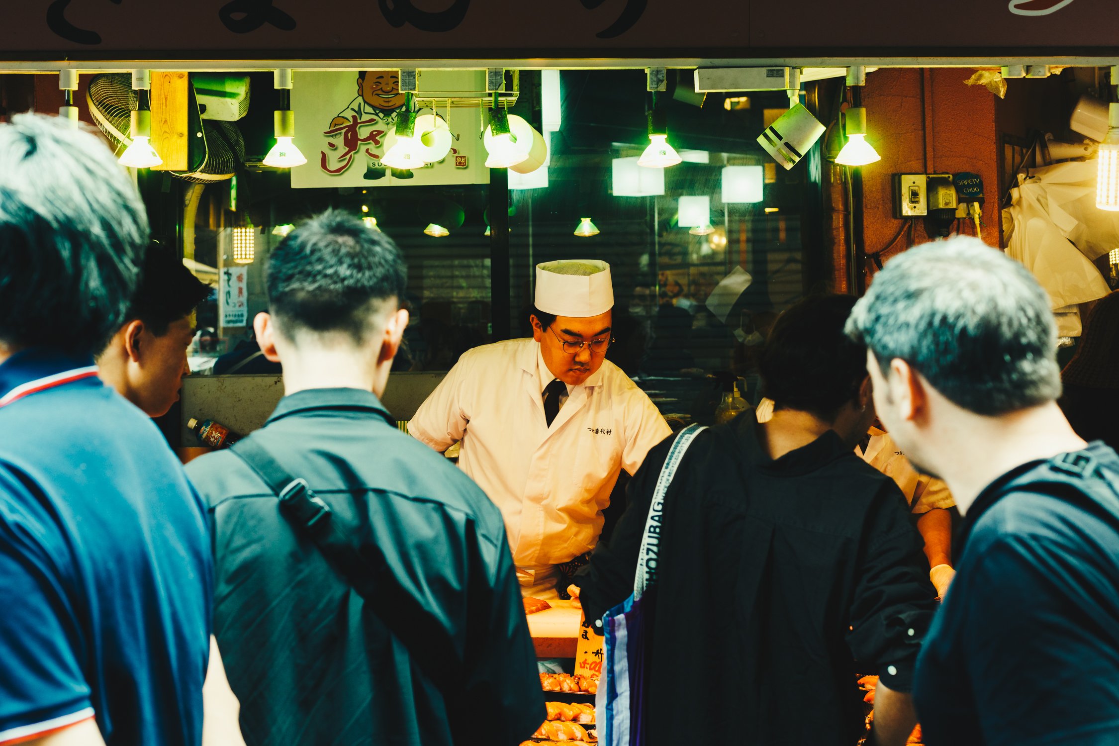A group of people gathered around a Japanese chef preparing sushi in a restaurant. The chef is wearing a white hat and uniform, and there are hanging lights and Japanese signs in the background.