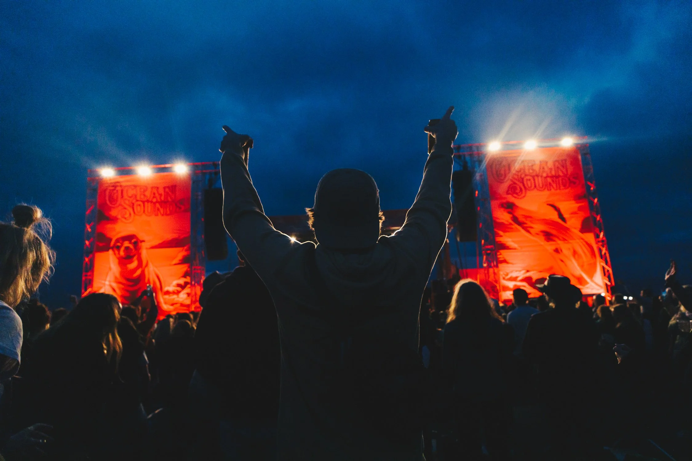 Person with arms raised at a concert or festival during evening, with large red screens displaying the words 'Urban Sounds' and imagery of a dog in the background, surrounded by an audience.