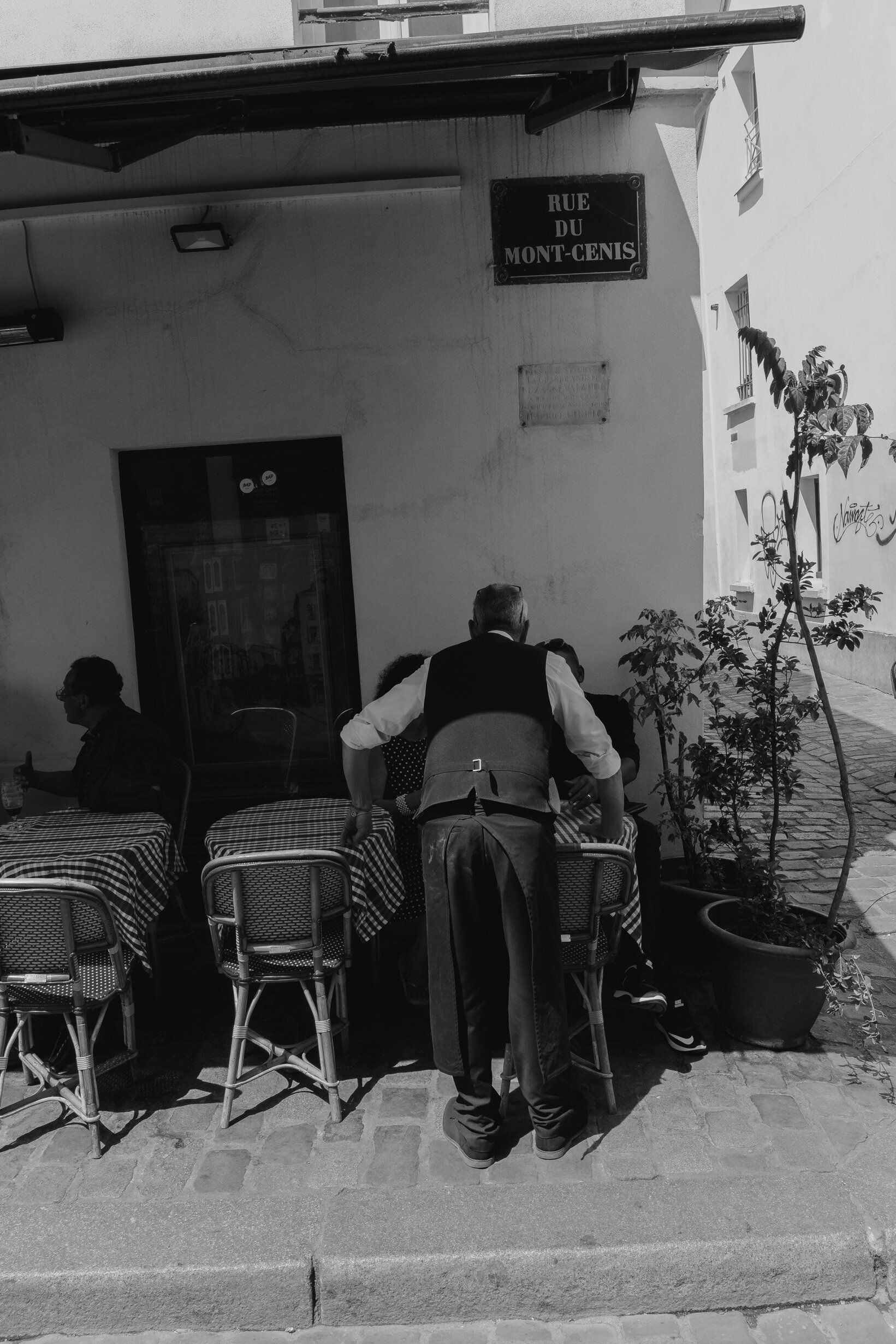 A man in a white shirt and vest leaning over a table with checkered tablecloths at an outdoor cafe. Another person is seated to his right, and a third person is on the left holding a drink. There are potted plants and a street sign that reads 'RUE DU